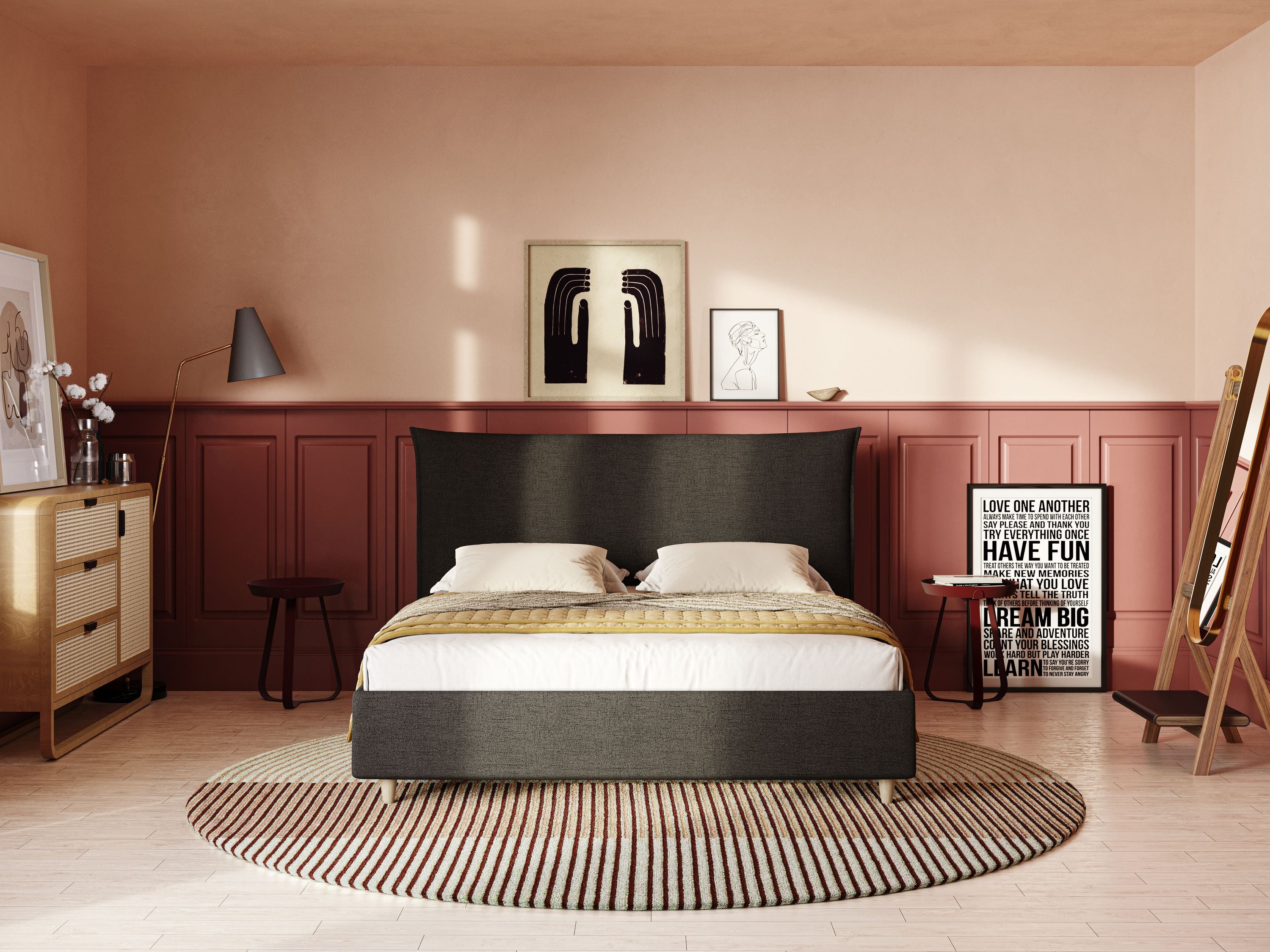 Una camera da letto moderna con il letto Audrey imbottito contenitore e sfoderabile in tessuto fiammato impermeabile T03 antracite di Letti.store, due cuscini, comodino in legno, opere d'arte a parete, tappeto a righe, comò in rattan, specchio, sgabello, citazione incorniciata e illuminazione calda.