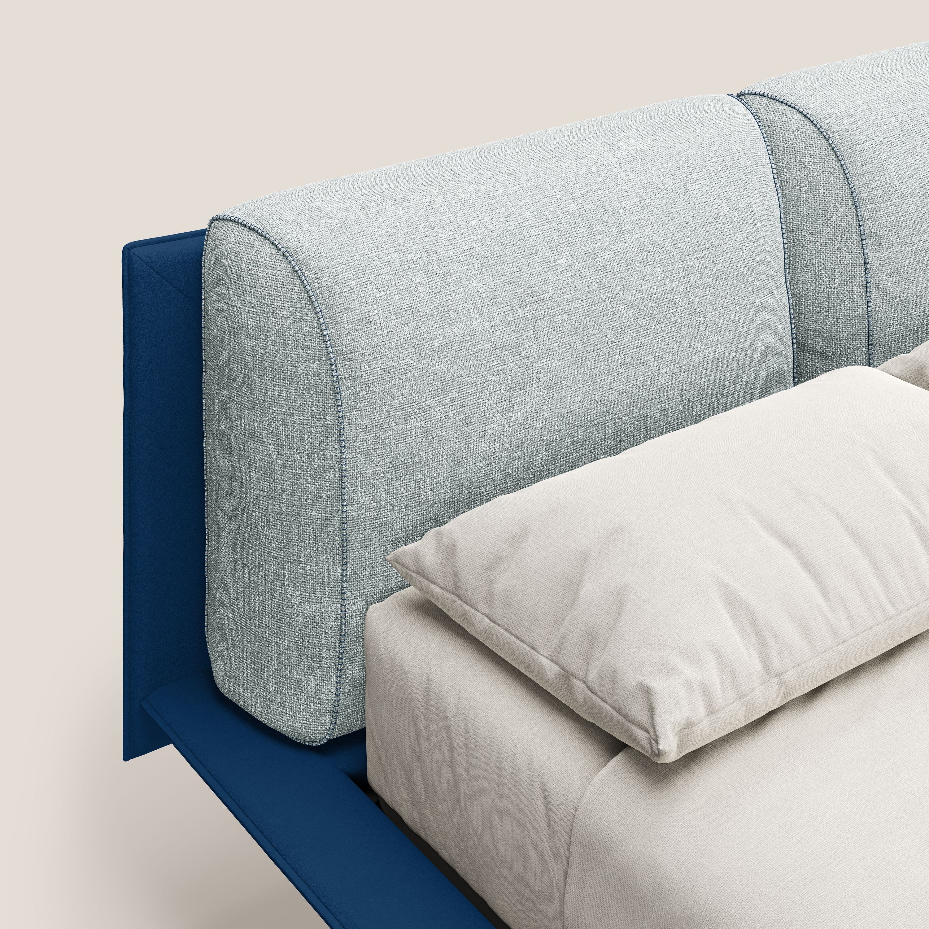 Un primo piano di Petra letto di design di Letti.store mostra la sua testata imbottita bicolore in tessuto T11 e T06 blu-ghiaccio, i cuscini in piuma, la biancheria da letto beige e la struttura blu intenso con piedi in mogano su uno sfondo chiaro.