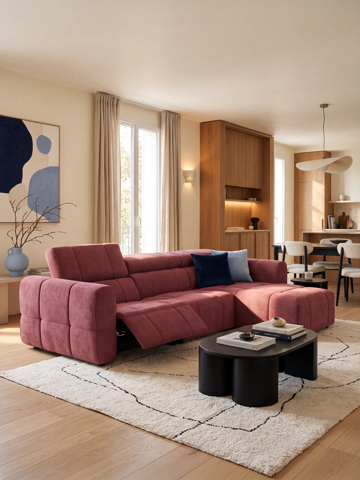 Un divano angolare modulare Giotto color rosa antico a 5 posti, lungo 296 cm, con penisola e funzione relax elettrica opzionale, rivestito in tessuto antimacchia T23, è posizionato su un tappeto color crema in una stanza inondata di luce solare; è dotato di chaise longue, poggiapiedi e un tavolino basso nero dalle forme organiche.