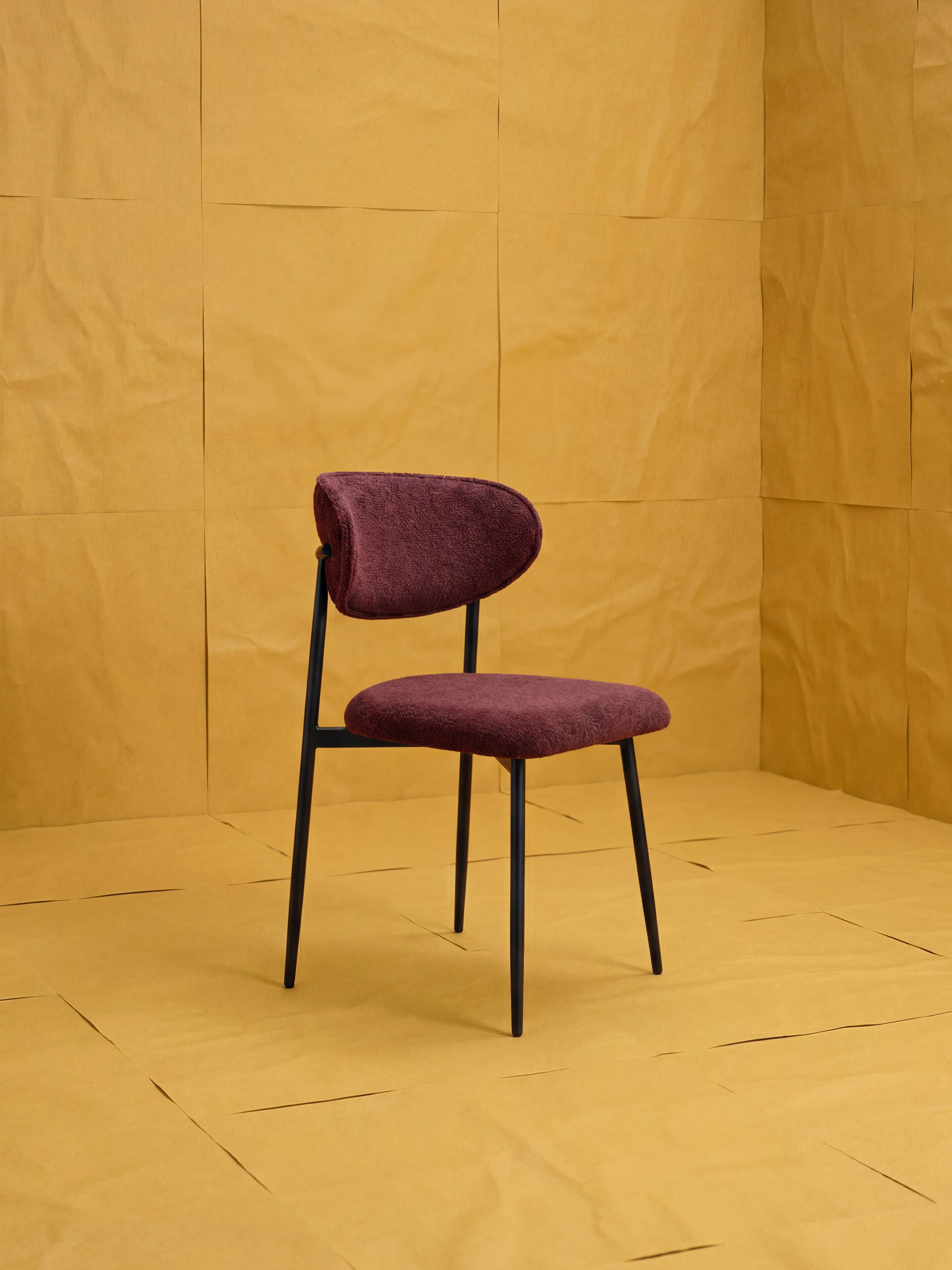 La sedia Luce di Divani.Store, imbottita in morbido tessuto ciniglia bordeaux, con schienale curvo e gambe in metallo nero, risalta su uno sfondo geometrico neutro: perfetta come sedia da cucina moderna.