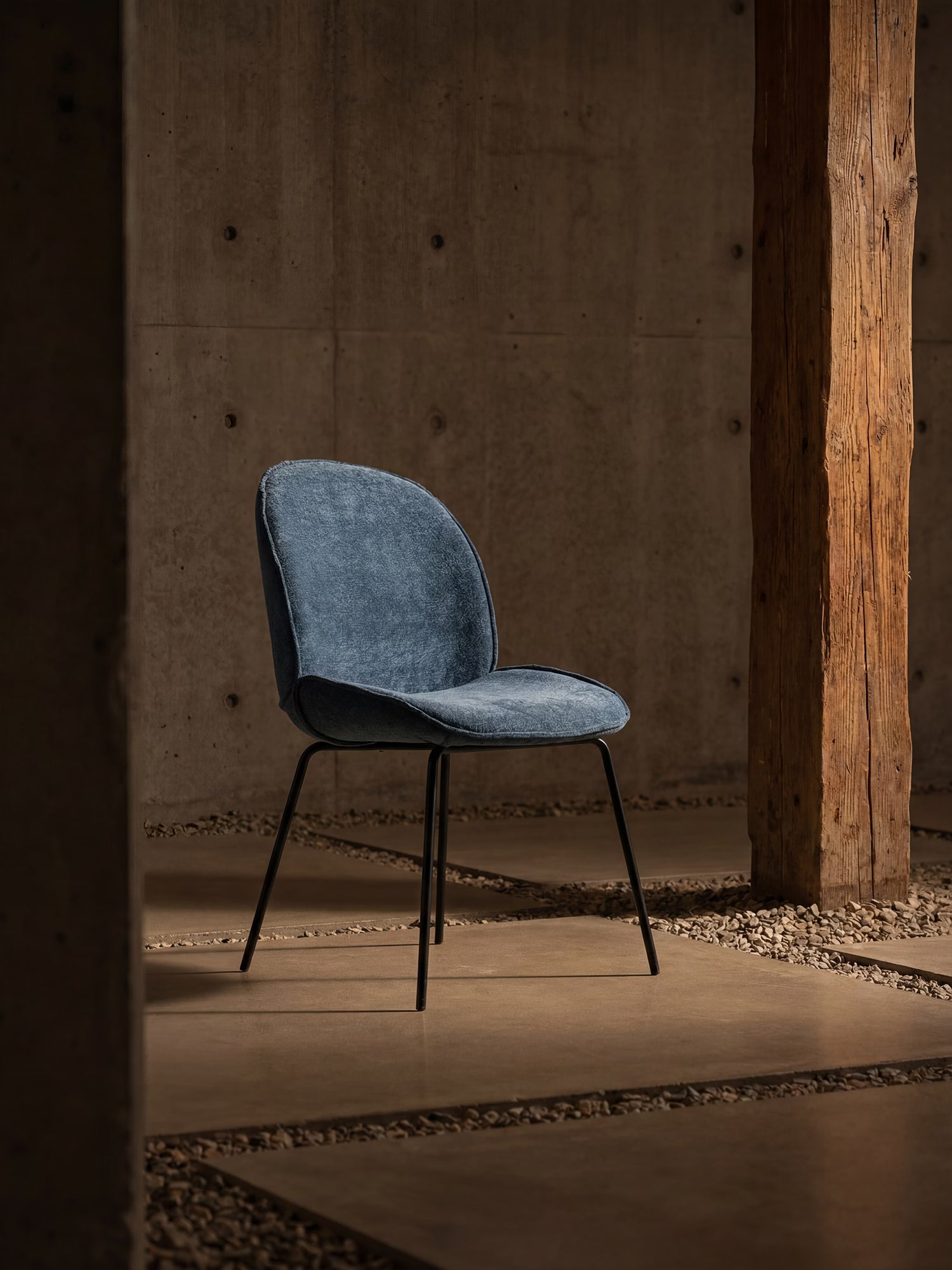 La sedia Iride, imbottita in morbido tessuto materico di Divani.Store, con gambe in metallo nero, si erge su una base di cemento lucido, circondata da pareti grigie, un alto palo di legno e piccole pietre sparse in un ambiente minimalista.