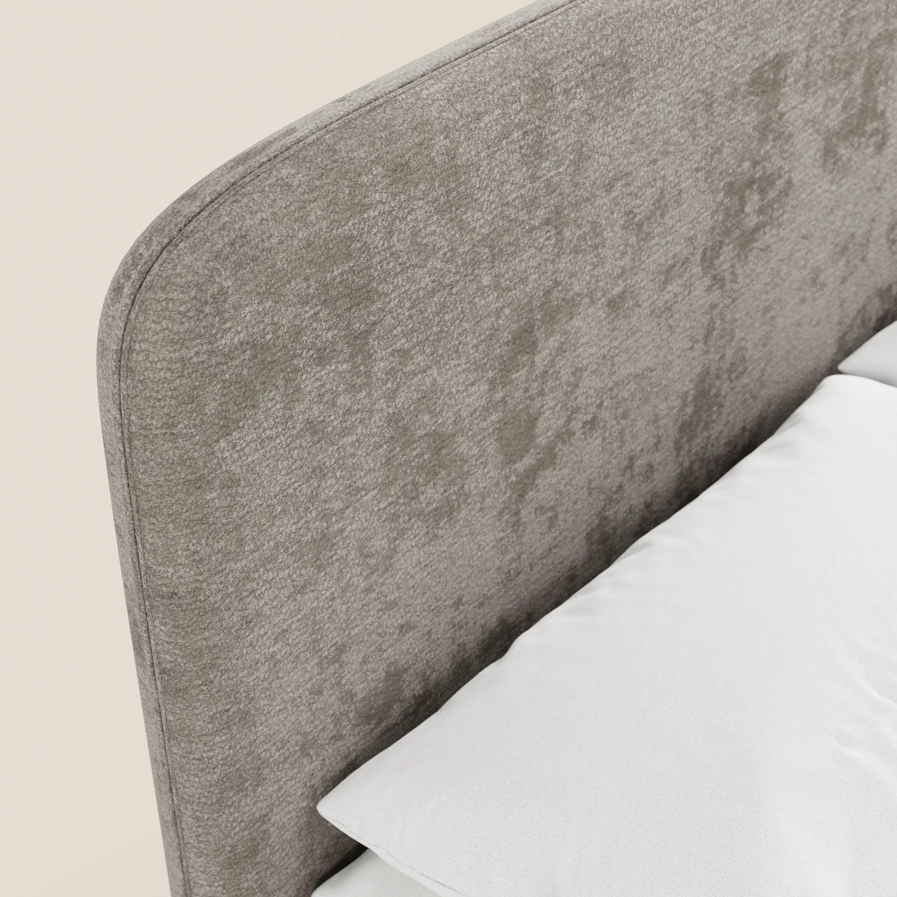 Un primo piano del letto imbottito KAELOS in stile minimal di Letti.store mette in risalto la sua testiera in ciniglia grigio chiaro, morbida e dalla trama particolare, con un cuscino bianco e la biancheria da letto in un angolo, il tutto su uno sfondo beige chiaro.