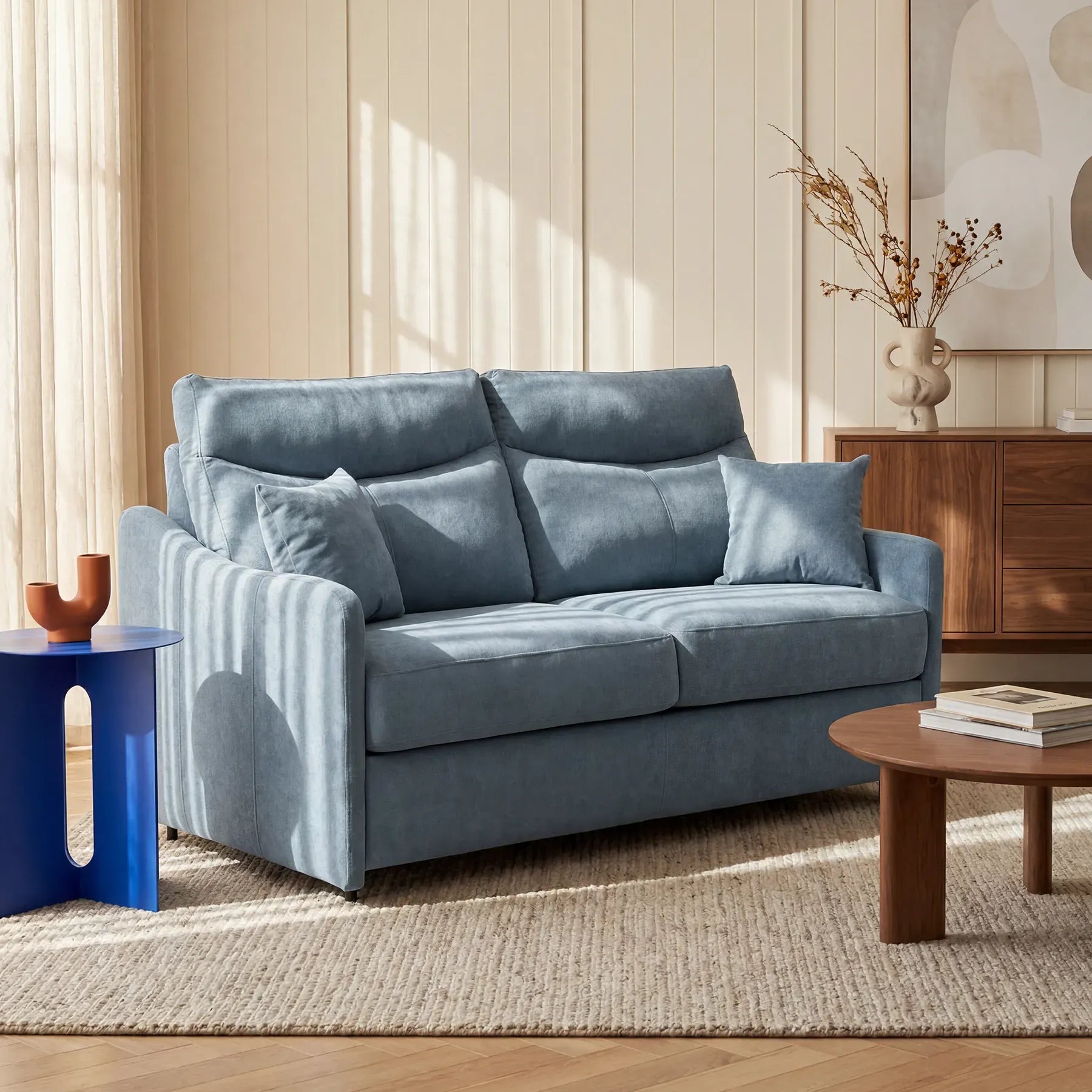 Un divano letto a tre posti Lodovico di colore azzurro, di Divani.Store, rivestito in tessuto antimacchia Oeko-Tex e dotato di un materasso alto 20 cm, poggia su un tappeto beige. A sinistra si trova un tavolino blu con un vaso in terracotta; davanti ad esso, un tavolino da salotto e alcuni libri; sopra la credenza è appesa un’opera d’arte astratta.