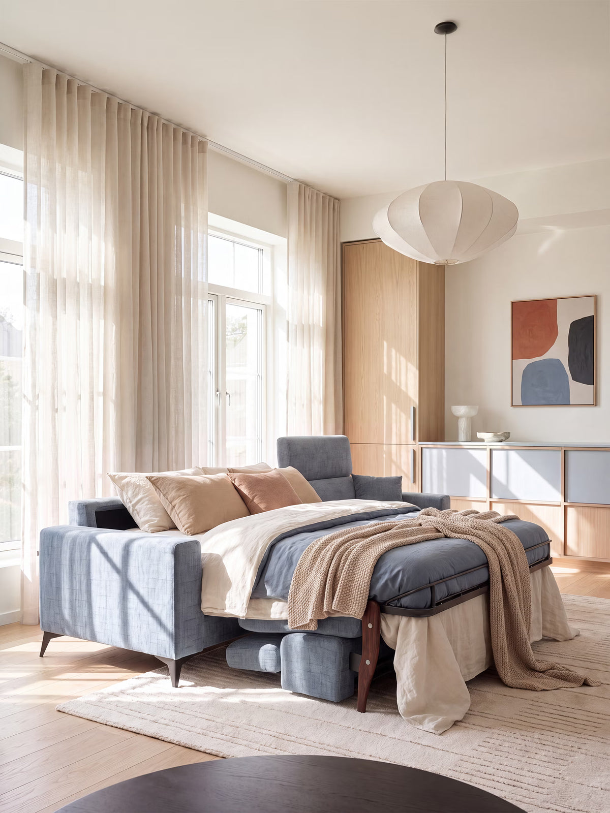 Una camera da letto inondata di luce ospita il divano letto angolare Pegaso con poggiatesta reclinabili e penisola contenitore in tessuto jacquard smacchiabile T22 di Divani.Store in una tonalità di grigio-blu, impreziosito da biancheria da letto nei toni del beige, del marrone chiaro e del blu, da un plaid in maglia e da morbidi cuscini. Tende color cuoio chiaro, pavimenti in legno, opere d’arte minimaliste e un moderno lampadario a sospensione completano l’arredamento accogliente e arioso.