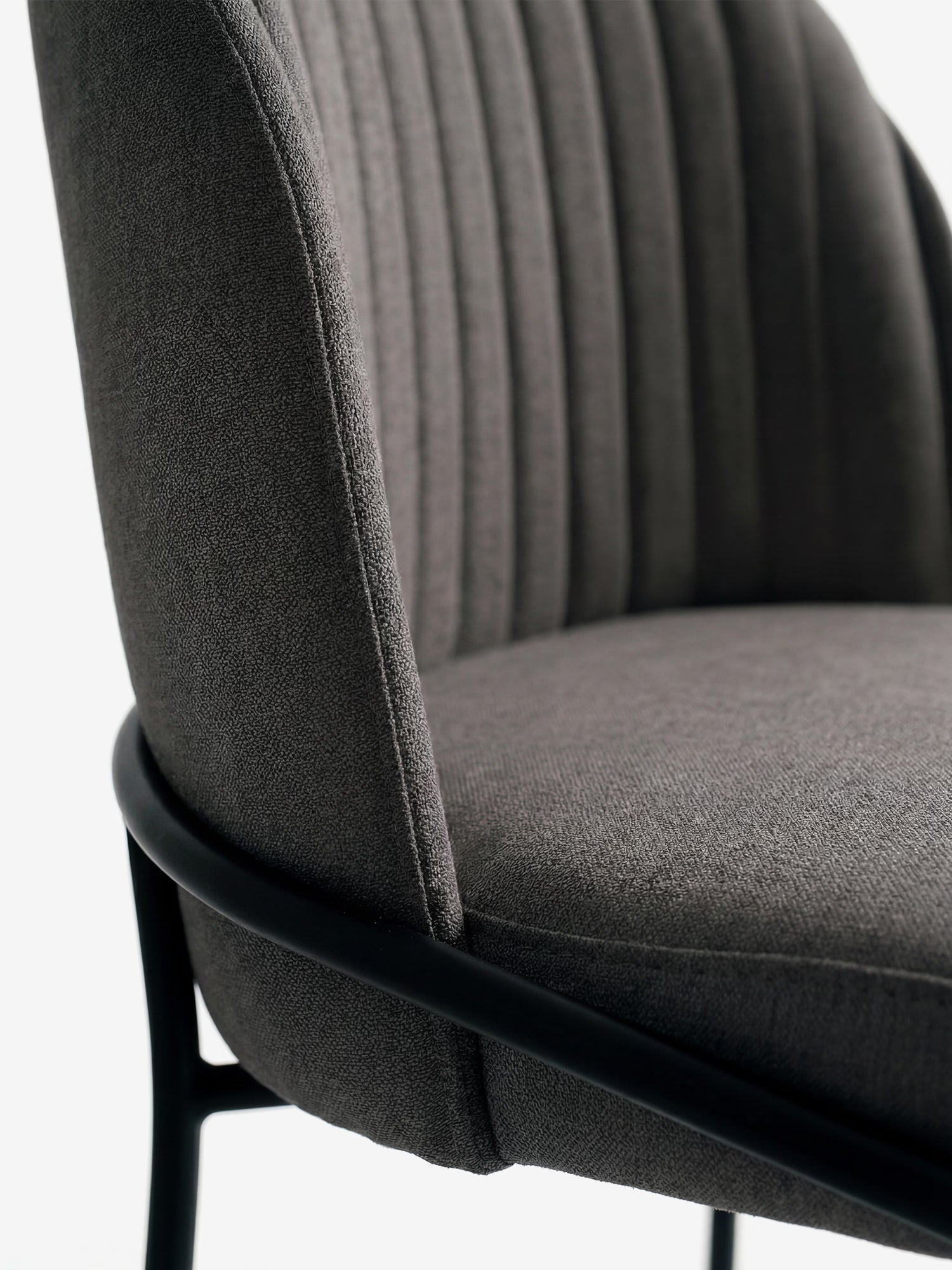 Primo piano della sedia imbottita "Plissè" in tessuto impermeabile di Divani.Store, caratterizzata da uno schienale plissettato, una seduta arrotondata e gambe in metallo nero opaco. La sedia è rivestita in tessuto grigio scuro. Sfondo bianco a tinta unita.