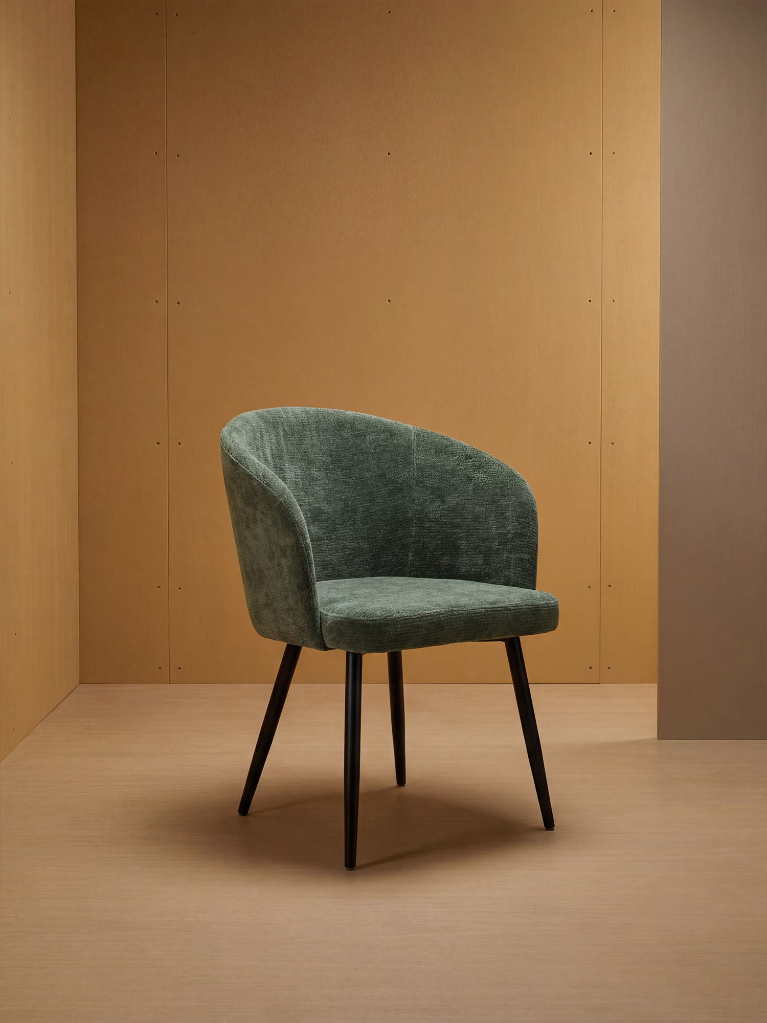 La sedia Rovo con braccioli di Divani.Store, caratterizzata da uno schienale curvo in ciniglia e da gambe in metallo nero, è esposta in un ambiente minimalista con pareti e pavimento in legno marrone chiaro che ne esaltano il design contemporaneo.