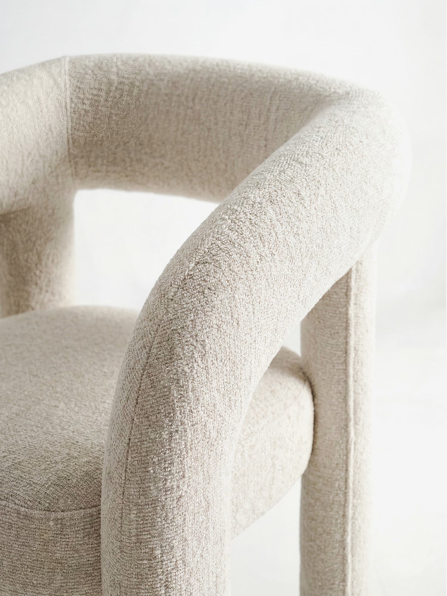 Un primo piano della sedia di design Sibilla di Divani.Store, caratterizzata da una forma arcuata e da una struttura interamente rivestita in morbido tessuto beige con braccioli e schienale arrotondati, su uno sfondo bianco a tinta unita.