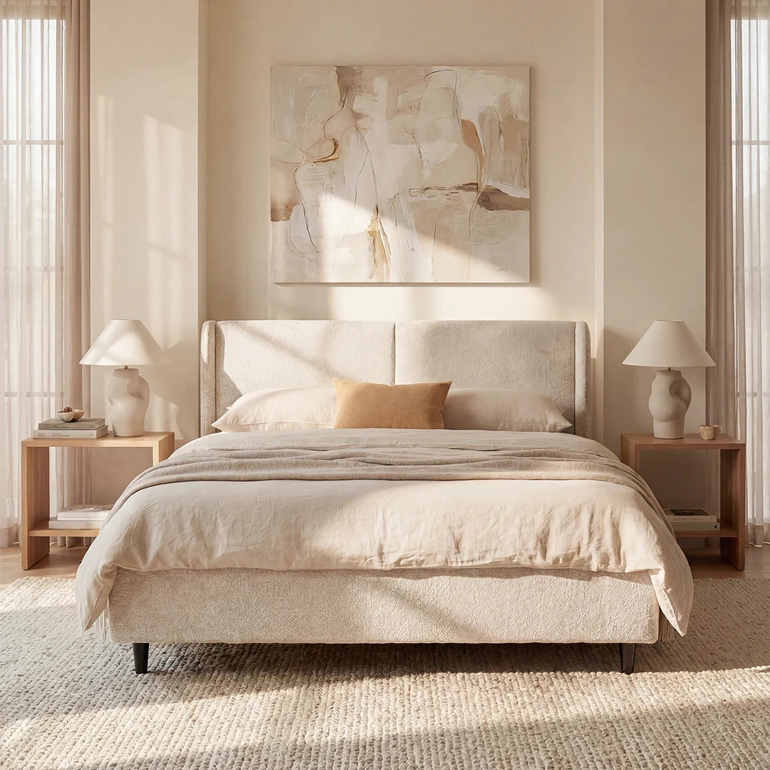 Una camera da letto dall'atmosfera serena è arredata con il letto di design VALKIRIA con contenitore e testiera avvolgente di Letti.store in beige chiaro, biancheria da letto color cuoio, un piccolo cuscino color cuoio, comodini in legno con lampade scultoree e libri, un'opera d'arte astratta appesa alla parete e la luce del sole che filtra attraverso le tende trasparenti.
