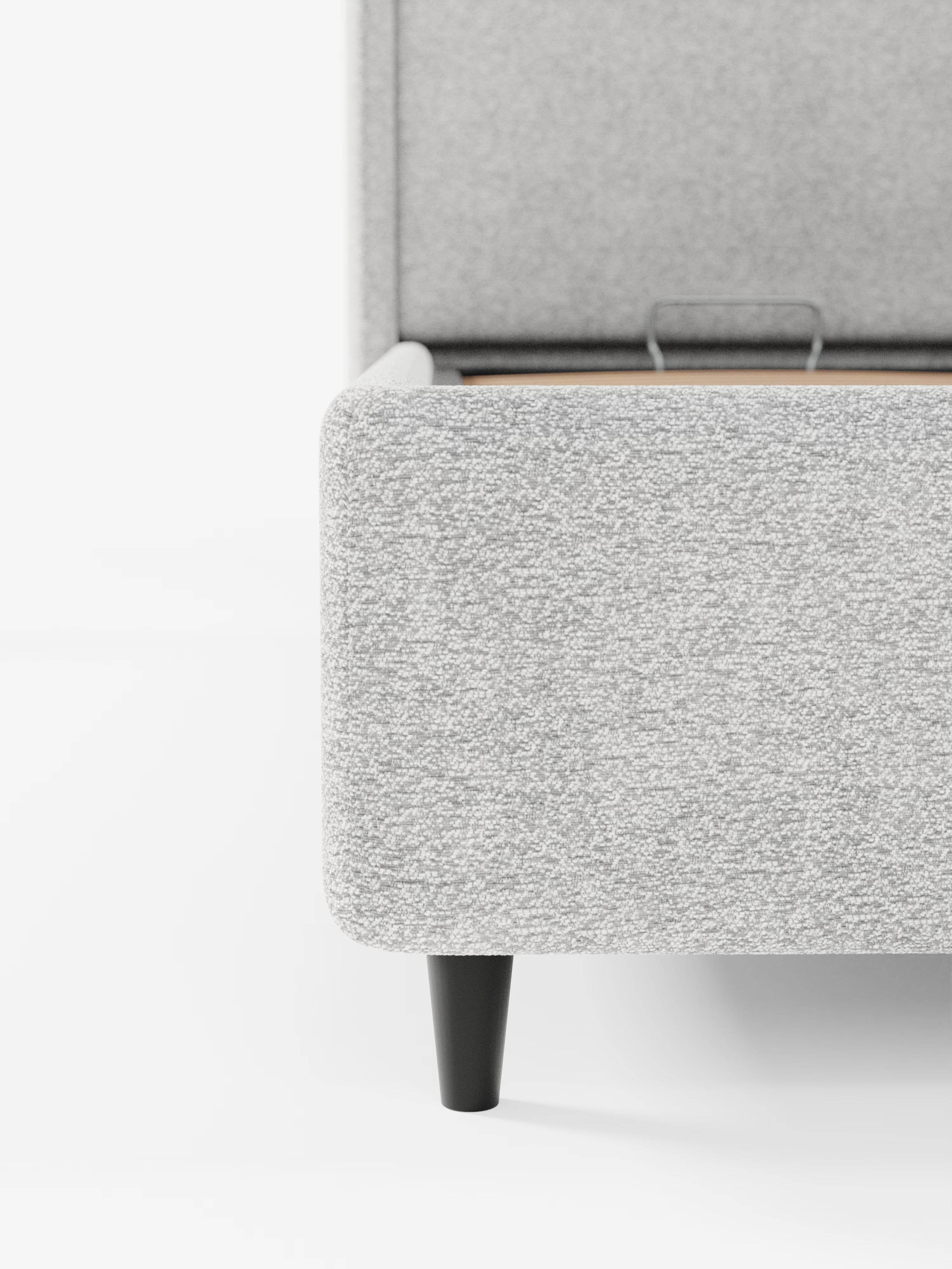 Primo piano dell'angolo di un letto di design VALKIRIA con vano contenitore e testiera avvolgente di Letti.store, rivestito in morbido tessuto Oeko-Tex® dalla trama ruvida, color grigio chiaro. La struttura contemporanea è caratterizzata da due cuscini imbottiti e poggia su eleganti gambe affusolate di colore nero. Le doghe del letto e una staffa di sostegno in metallo sono parzialmente visibili su uno sfondo bianco a tinta unita.