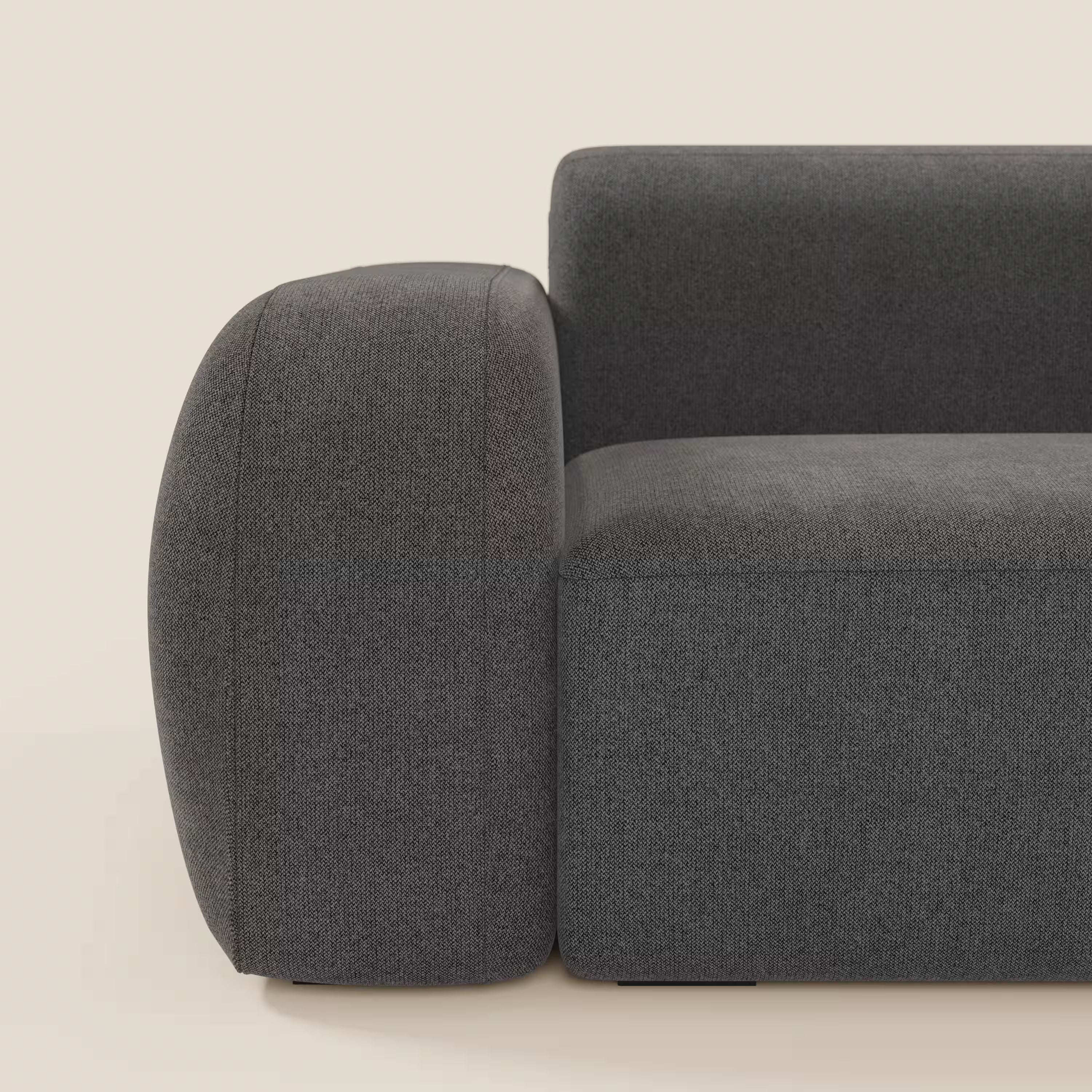 Primo piano del BonBon Divano modulare Lineare 220 di Divani.Store in tessuto smacchiabile T23 antracite, che mostra i moderni bordi arrotondati e gli spessi braccioli su uno sfondo beige chiaro. È visibile solo una parte del divano.