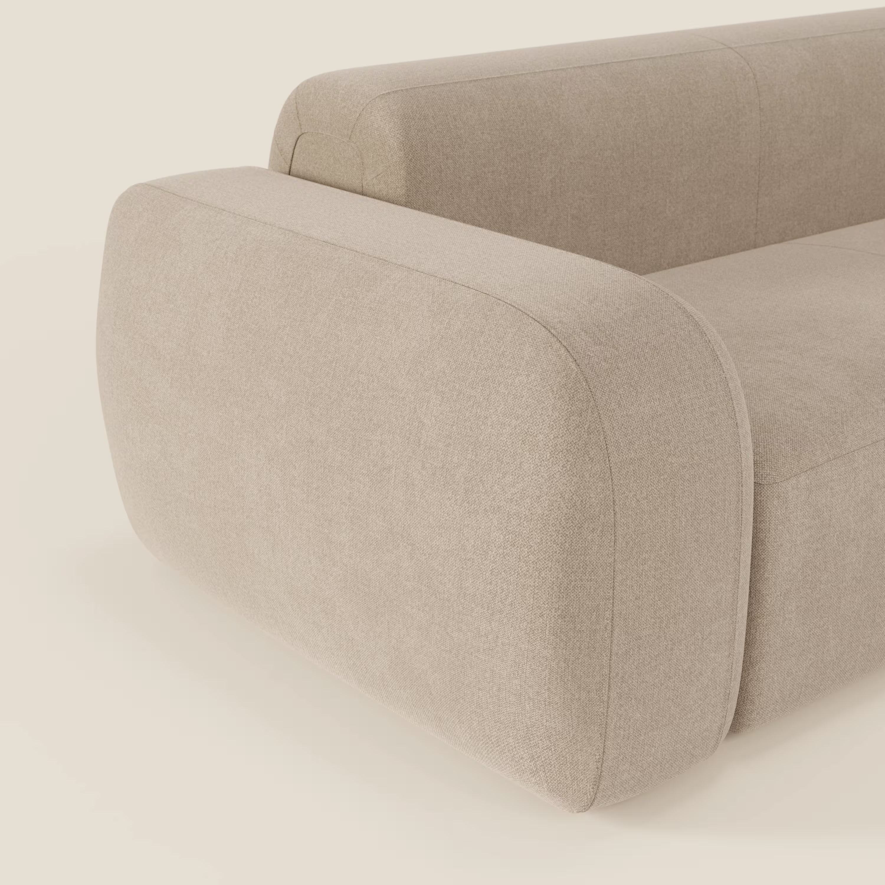 Primo piano del BonBon Divano componibile di Divani.Store in tessuto smacchiabile T23 beige, caratterizzato da braccioli spessi e arrotondati e da un design minimalista. Sullo sfondo crema è visibile solo una parte del divano con pouf terminale.