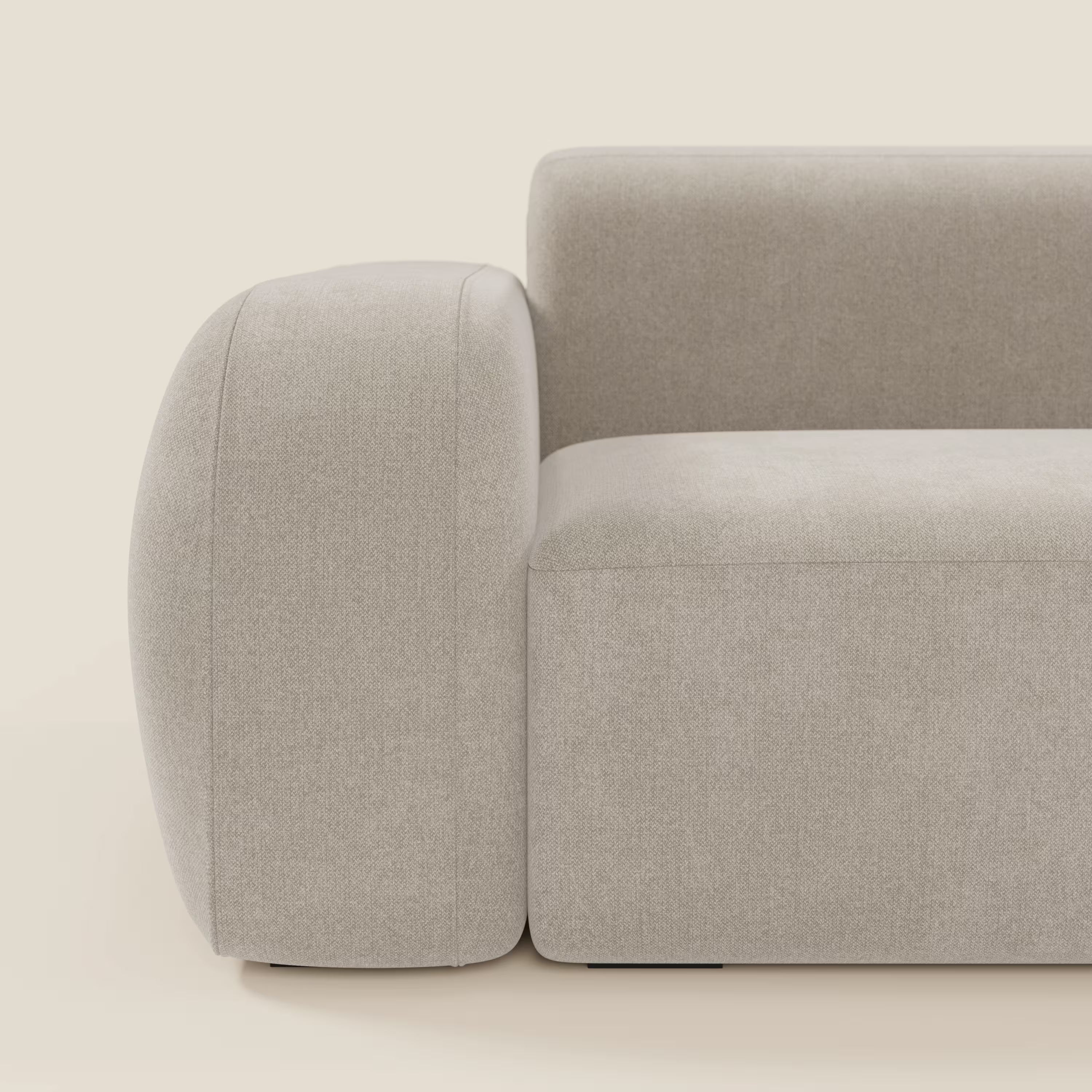 Un primo piano di BonBon Divano componibile di Divani.Store in tessuto smacchiabile T23 panna mostra l'ampio bracciolo sinistro arrotondato che si fonde con la seduta rettangolare. Il design minimalista emana un comfort soffice su uno sfondo bianco sporco.