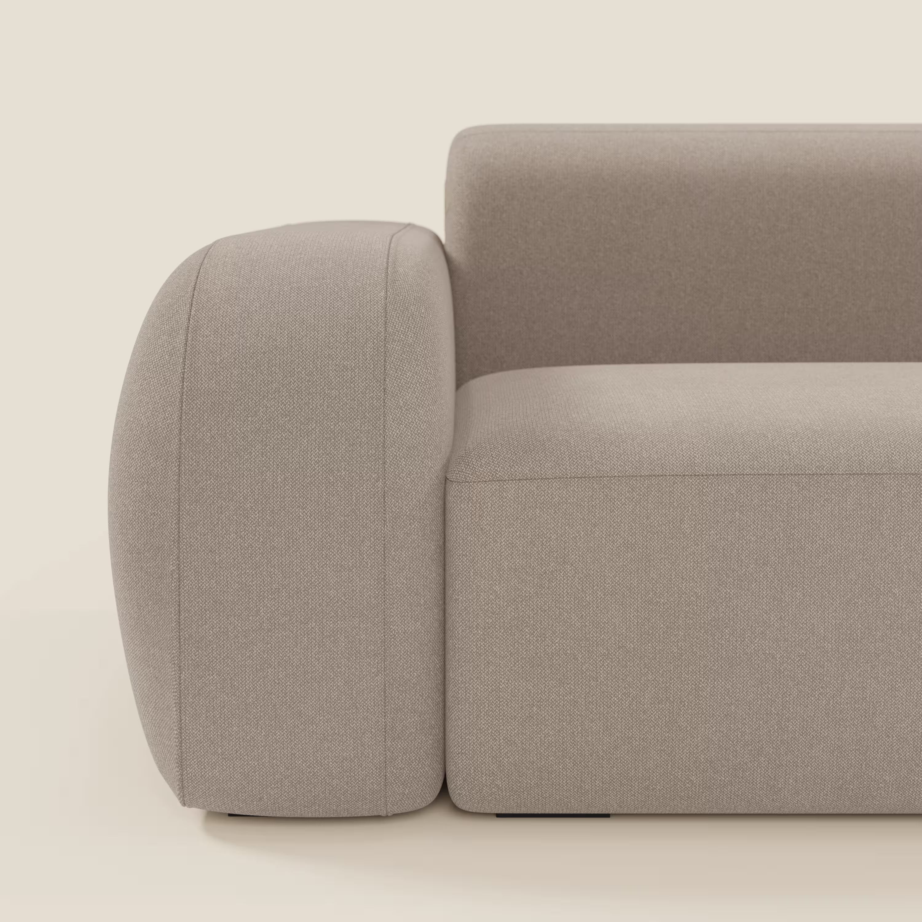 Un primo piano del divano componibile BonBon Divano di Divani.Store in smacchiabile tortora T23, caratterizzato da braccioli spessi e arrotondati e da un pouf terminale, mette in evidenza il suo moderno design geometrico su uno sfondo bianco sporco.