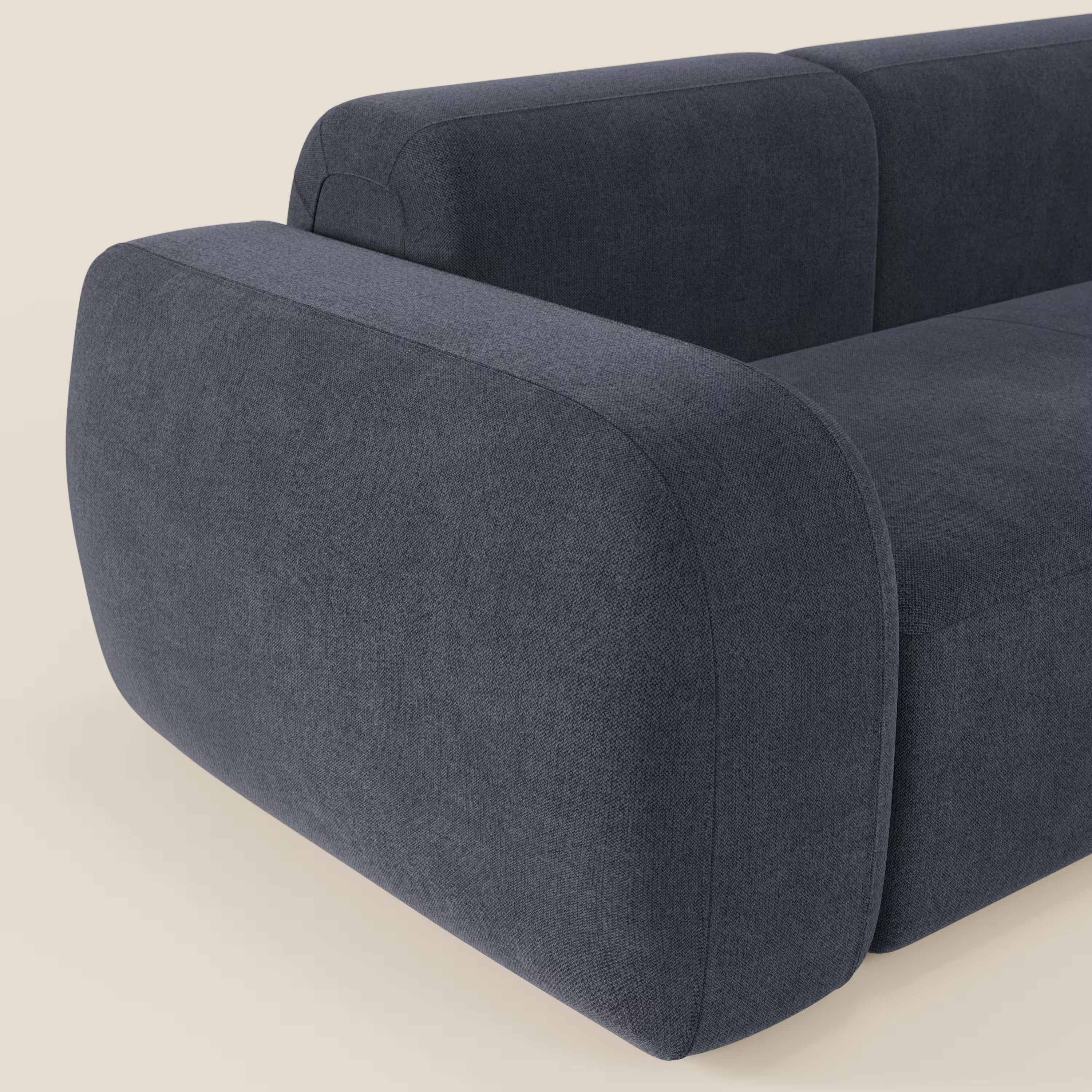 Un primo piano del BonBon Divano modulare componibile - Composizione Spaccamattone di Divani.Store in smacchiabile T23 blu ardesia mostra i braccioli spessi e arrotondati e i cuscini in morbida texture, sullo sfondo di un semplice beige. È visibile solo una parte del divano.