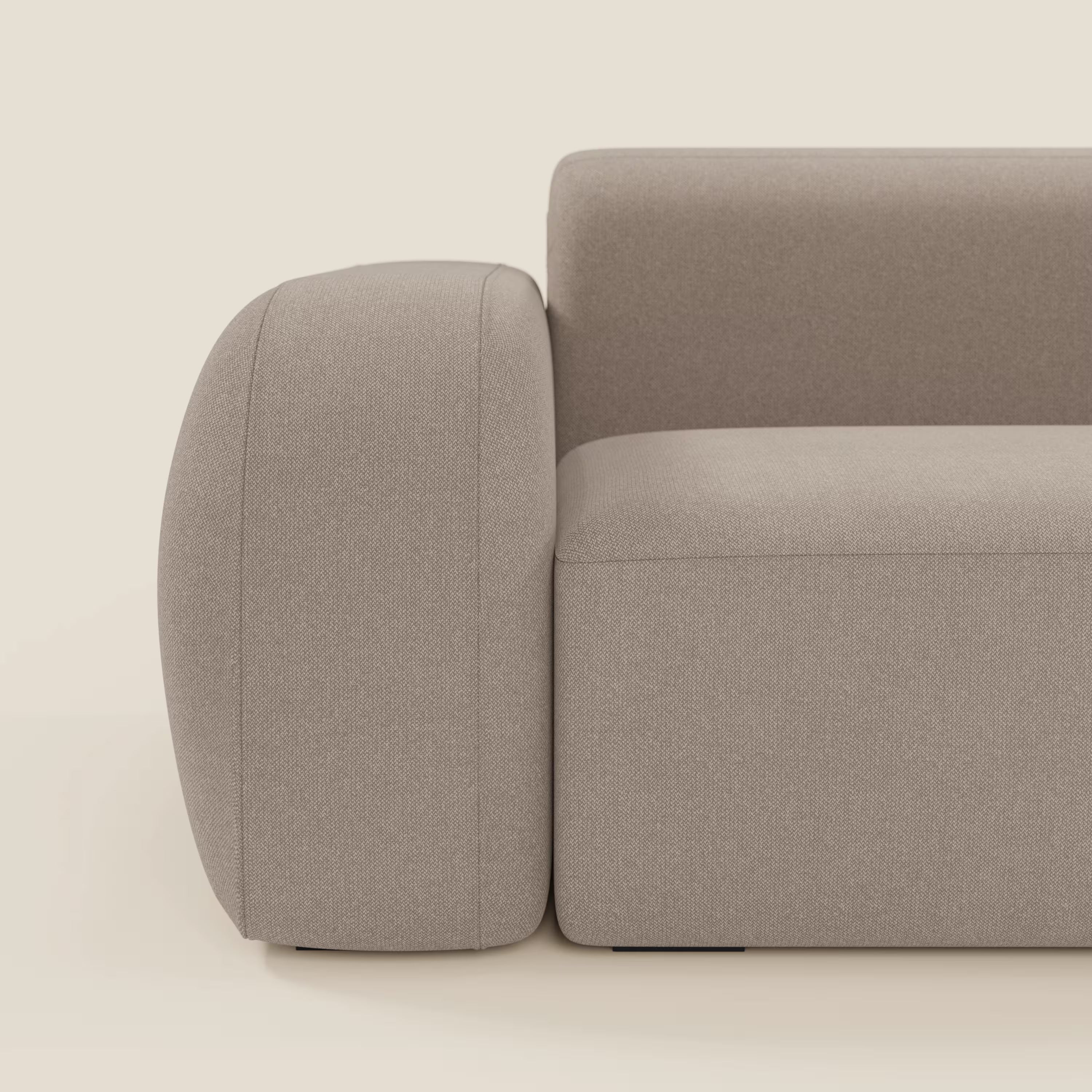 Primo piano del divano angolare componibile e modulare BonBon di Divani.Store in tessuto smacchiabile T23 tortora, che mostra il bracciolo sinistro, la seduta e lo schienale con bordi lisci e arrotondati su uno sfondo crema chiaro.
