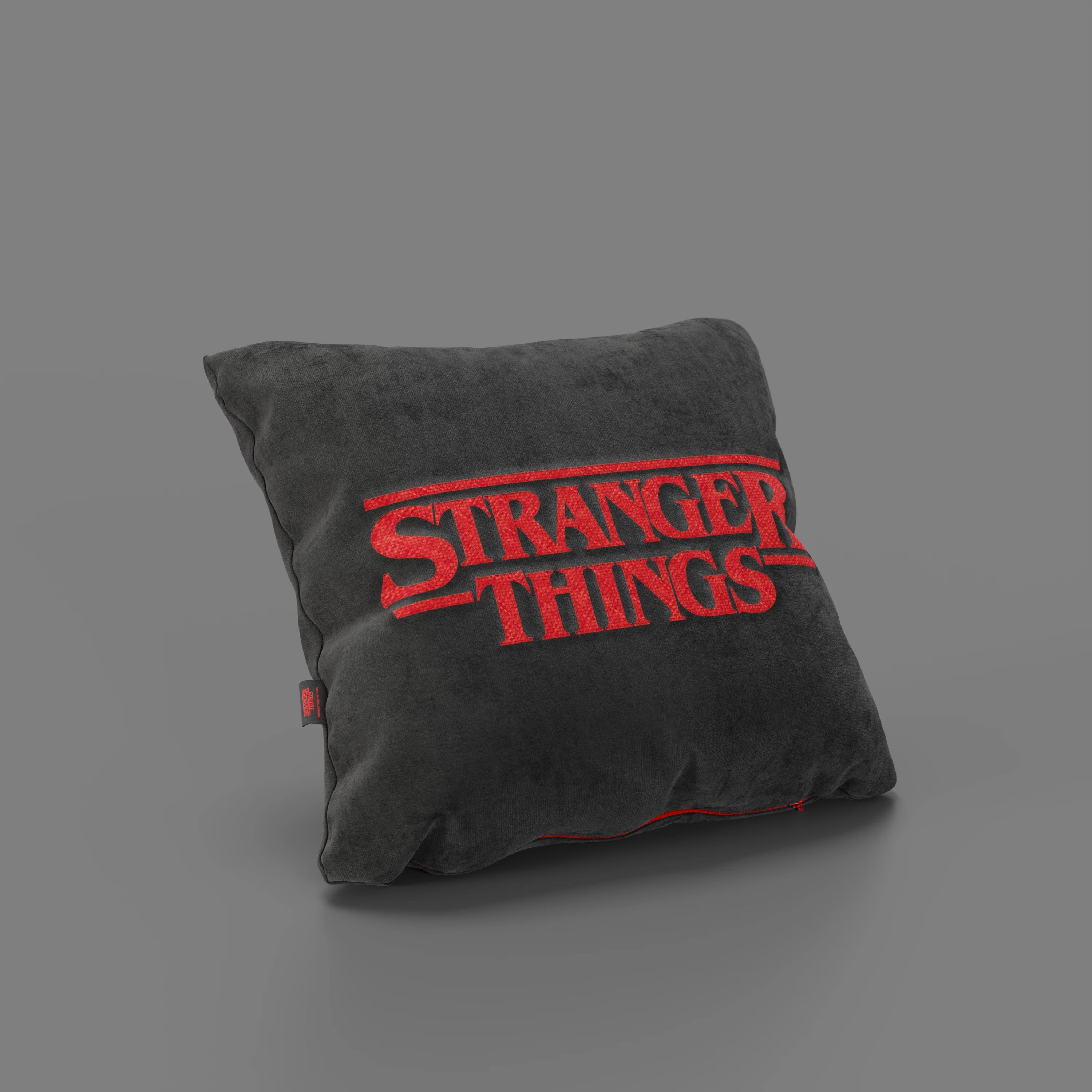 Il Cuscino Stranger Things Limited Edition di Divani.Store, di forma quadrata e di colore antracite, poggia su una superficie grigia. Il cuscino, resistente alle macchie, presenta un audace ricamo rosso del logo di "Stranger Things" e dettagliate cuciture rosse.