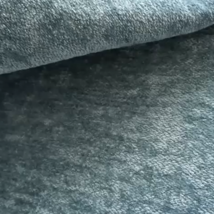 Un primo piano di due strati di T27 Brezza di Divani.Store mostra la sua superficie goffrata, morbida ed elegante, con una sensazione di peluche e un sottile aspetto marmorizzato. Il tessuto grigio-blu, resistente alle macchie, è delicatamente piegato per evidenziare la sua consistenza vellutata.