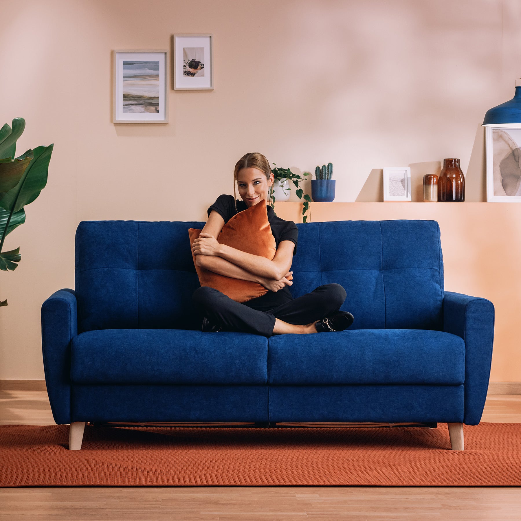 Una donna siede a gambe incrociate sul Callisto Divano letto matrimoniale in tessuto morbido impermeabile T02 blu di Divani.Store, abbracciata a un cuscino marrone. Pareti color pesca chiaro, opere d'arte incorniciate, piante, vasi e una lampada a sospensione blu decorano la stanza.