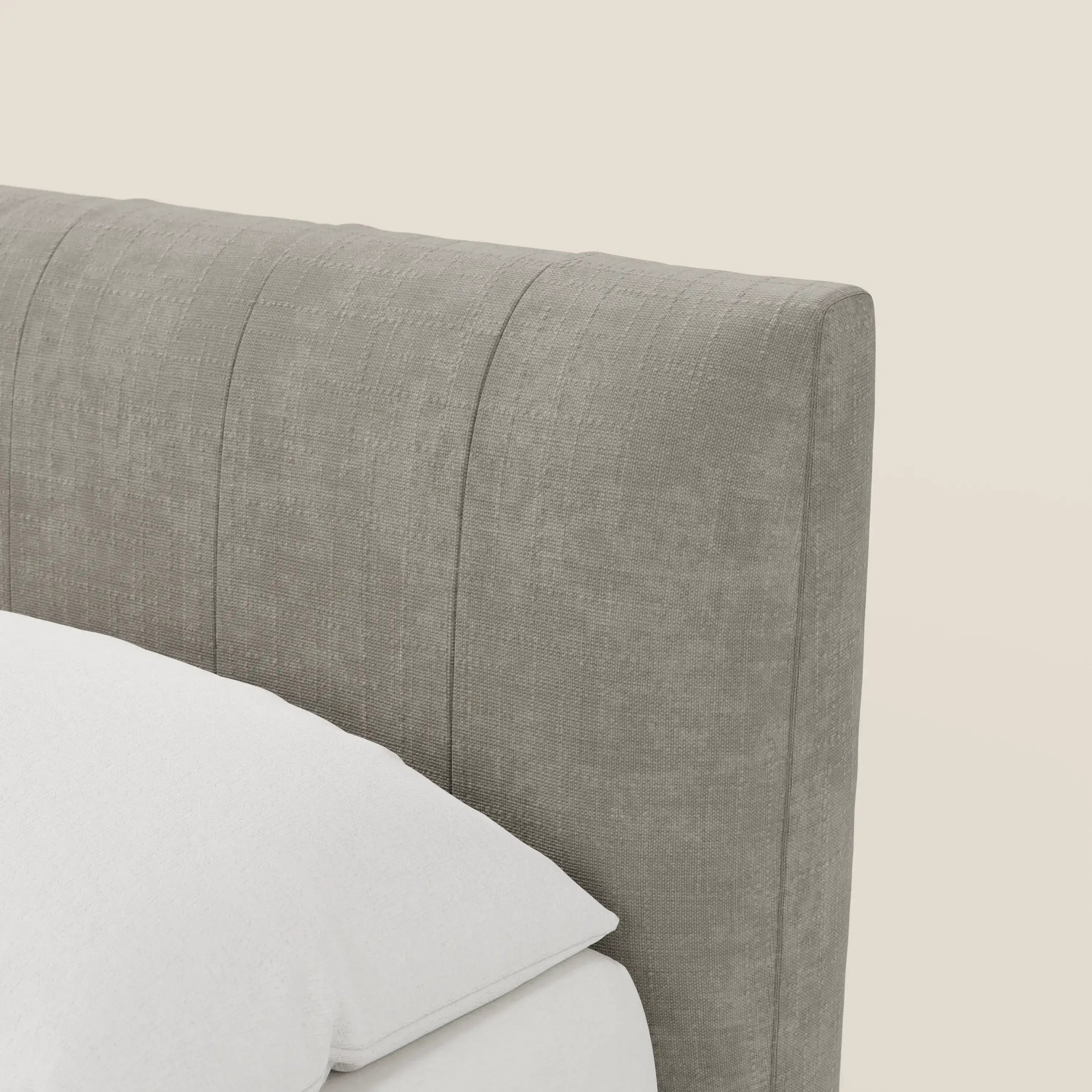 Primo piano della testiera del letto contenitore Letti.store Bogart Edizione Grande Fratello, rivestita in tessuto sfoderabile antimacchia T22 grigio chiaro con cuciture verticali. Un cuscino e un materasso bianchi appaiono in primo piano su uno sfondo beige.