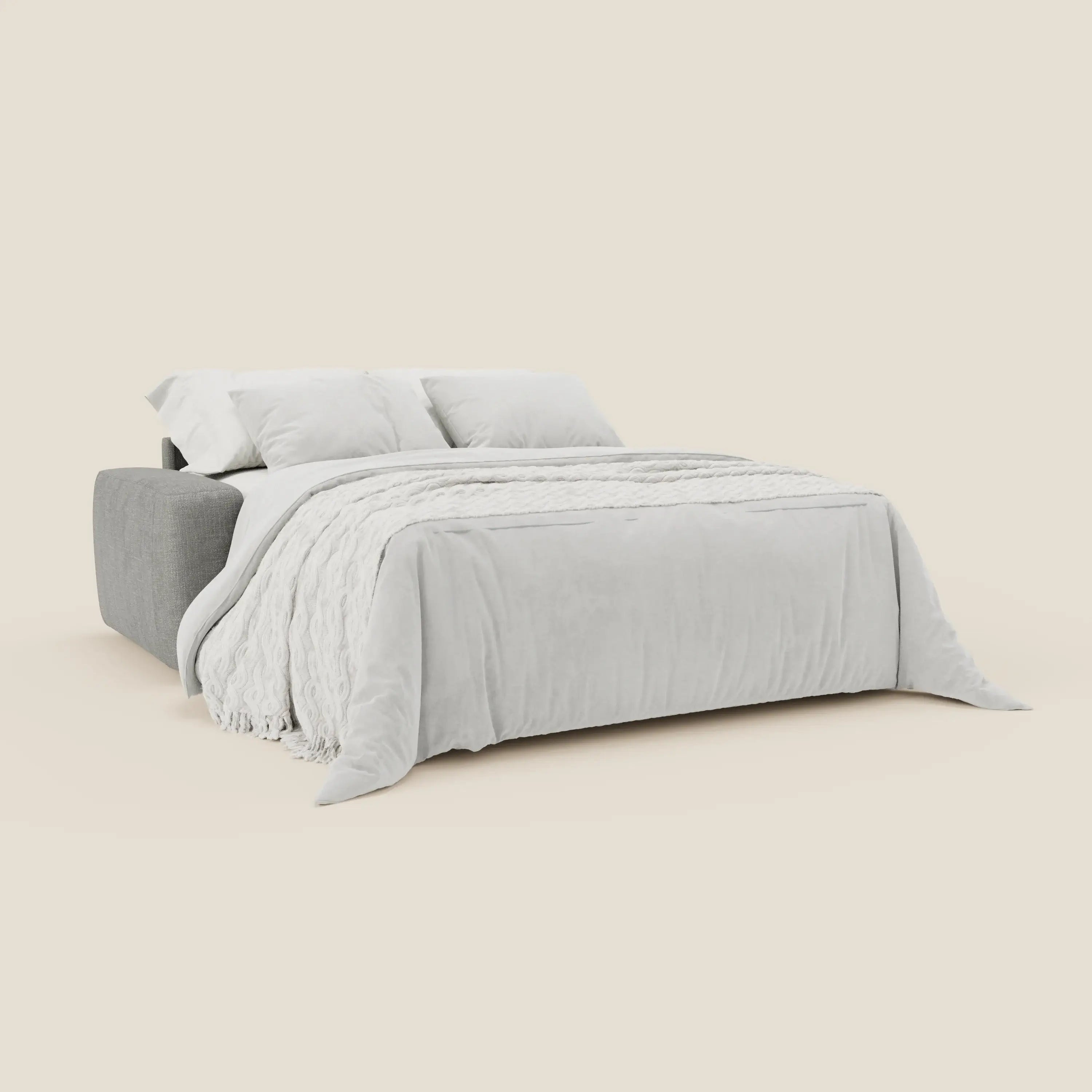 Il letto materasso Cloud Divano H17 cm di Divani.Store, in tessuto impermeabile grigio T06, si dispiega in un letto con una coperta bianca e tre cuscini bianchi. Un getto strutturato fa capolino, mentre lo sfondo beige ne sottolinea il design minimalista.
