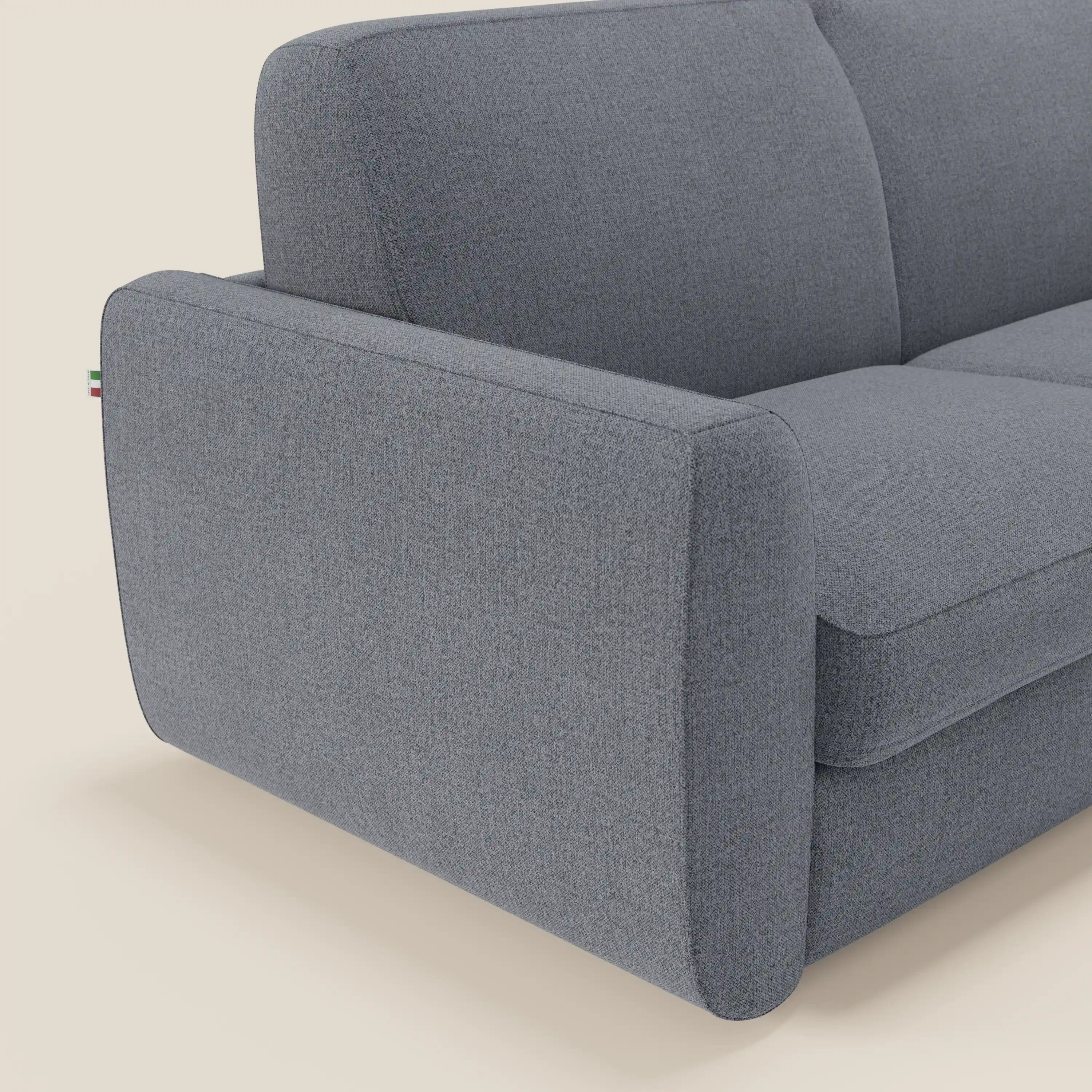 Un primo piano del divano letto Leonida sfoderabile e materasso H18 cm di Divani.Store in smacchiabile T23 blu grafite mostra gli ampi braccioli, i cuscini in peluche, la texture morbida e la targhetta della bandiera italiana sulla cucitura esterna.