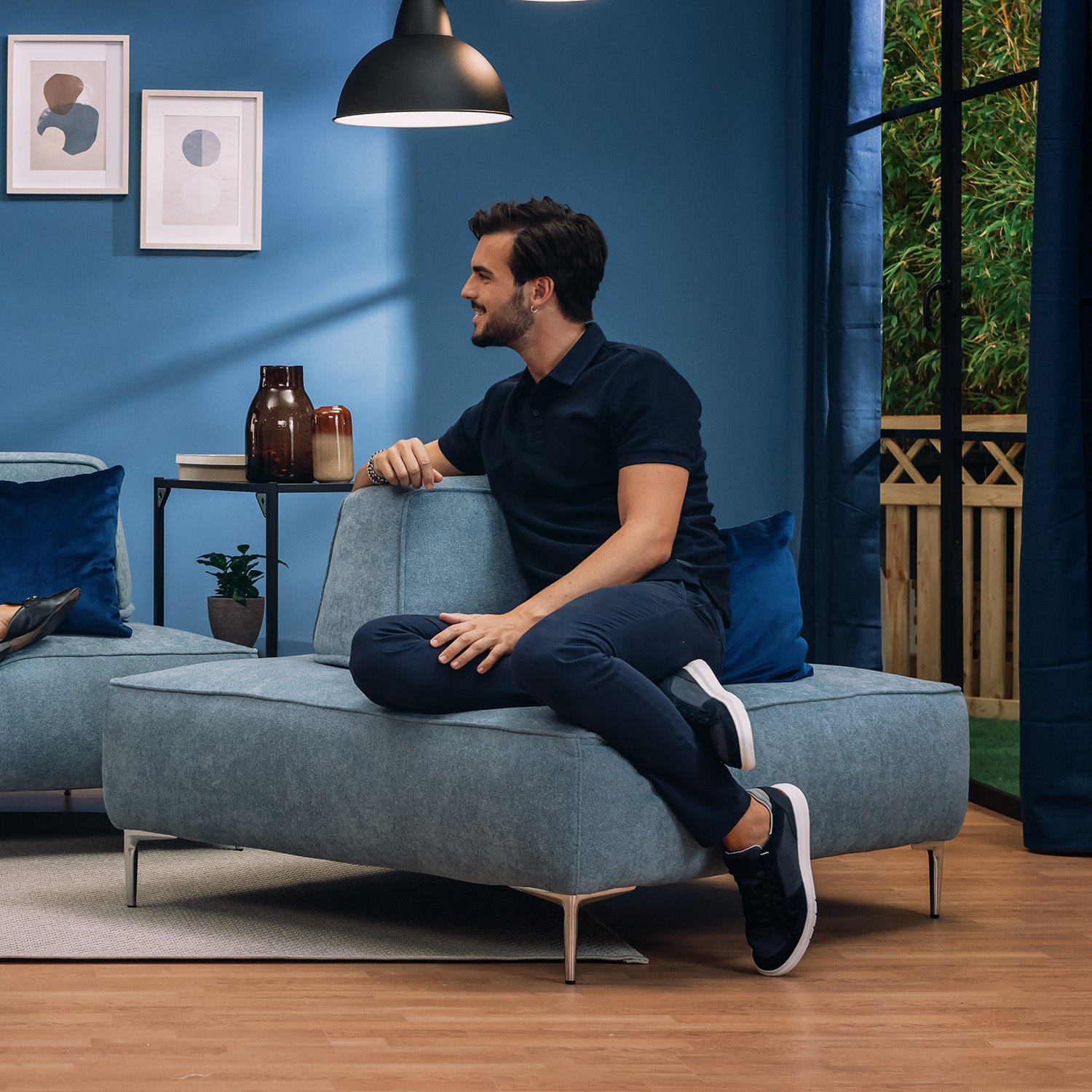 Un uomo con capelli corti e barba siede di lato sulla poltrona modulare Divani.Store Newton con schienali mobili T07 carta da zucchero in un soggiorno moderno con pareti blu, piante, vasi, arte e una finestra, sorridendo e guardando a sinistra.