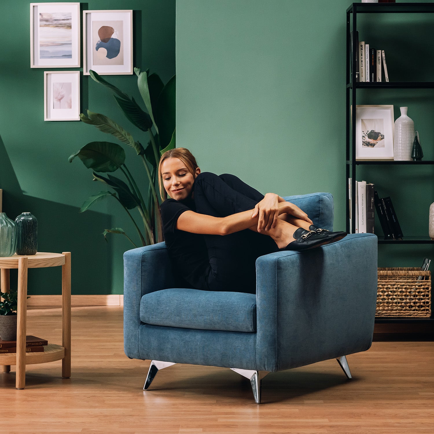Una donna vestita di nero siede soddisfatta su una poltroncina Divani.Store Orione in morbido tessuto impermeabile ad intreccio T06 azzurro, sorridendo ad occhi chiusi in un soggiorno moderno con pareti verdi, pavimento in legno e arredamento elegante.