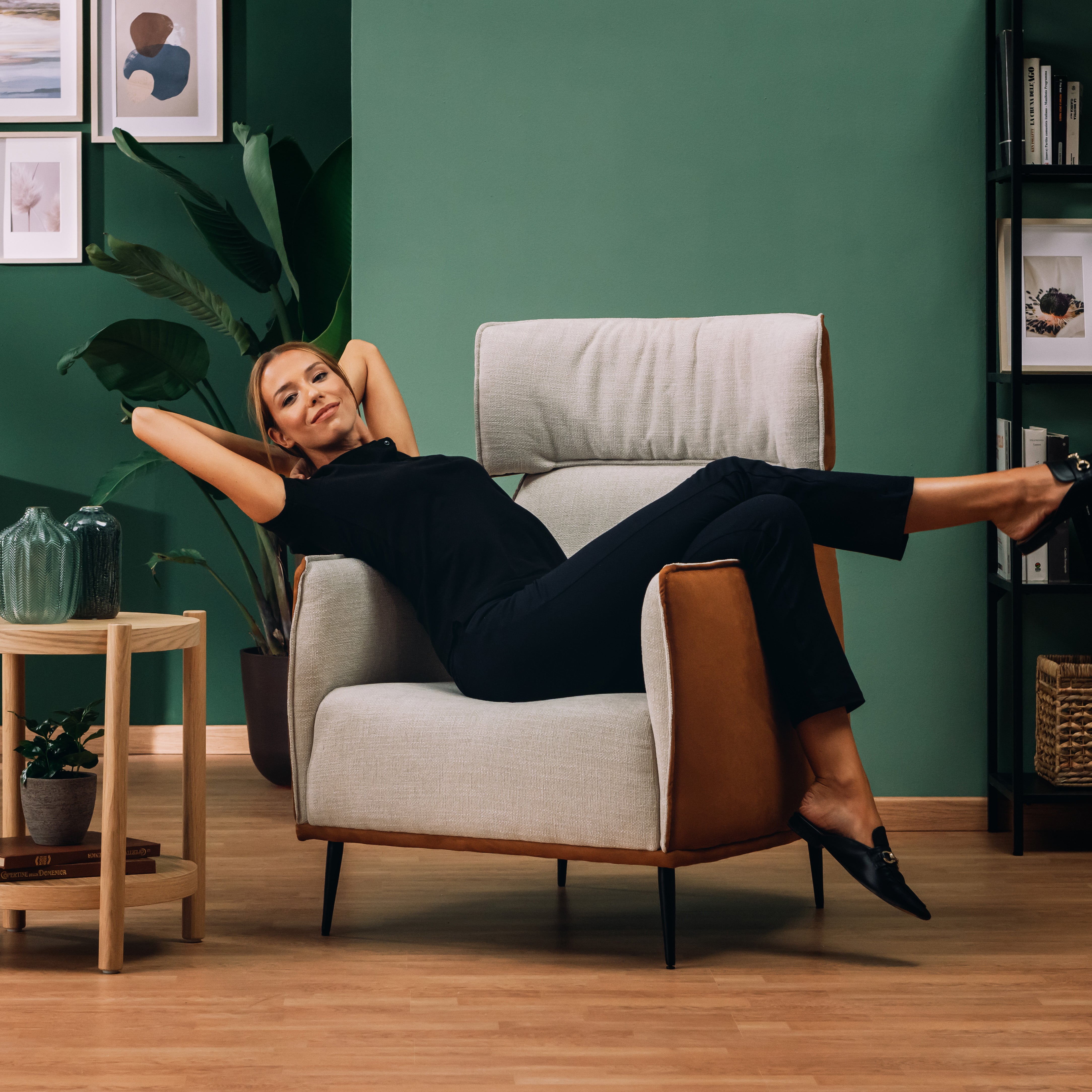 Una donna in abito nero si sdraia lateralmente sulla Zelda Poltroncina dal design moderno in morbido tessuto impermeabile T06 panna di Divani.Store, in un elegante salotto con parete verde, pavimento in legno, pianta in vaso, opere d'arte incorniciate e libreria.