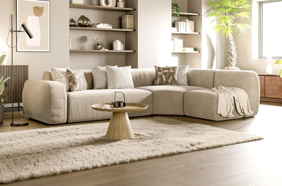 Un soggiorno moderno con divano componibile beige, cuscini neutri, tavolino in legno, mensole a muro con decorazioni, tappeto, pianta alta, grande finestra e travi a vista sul soffitto. Un'atmosfera calda e invitante.