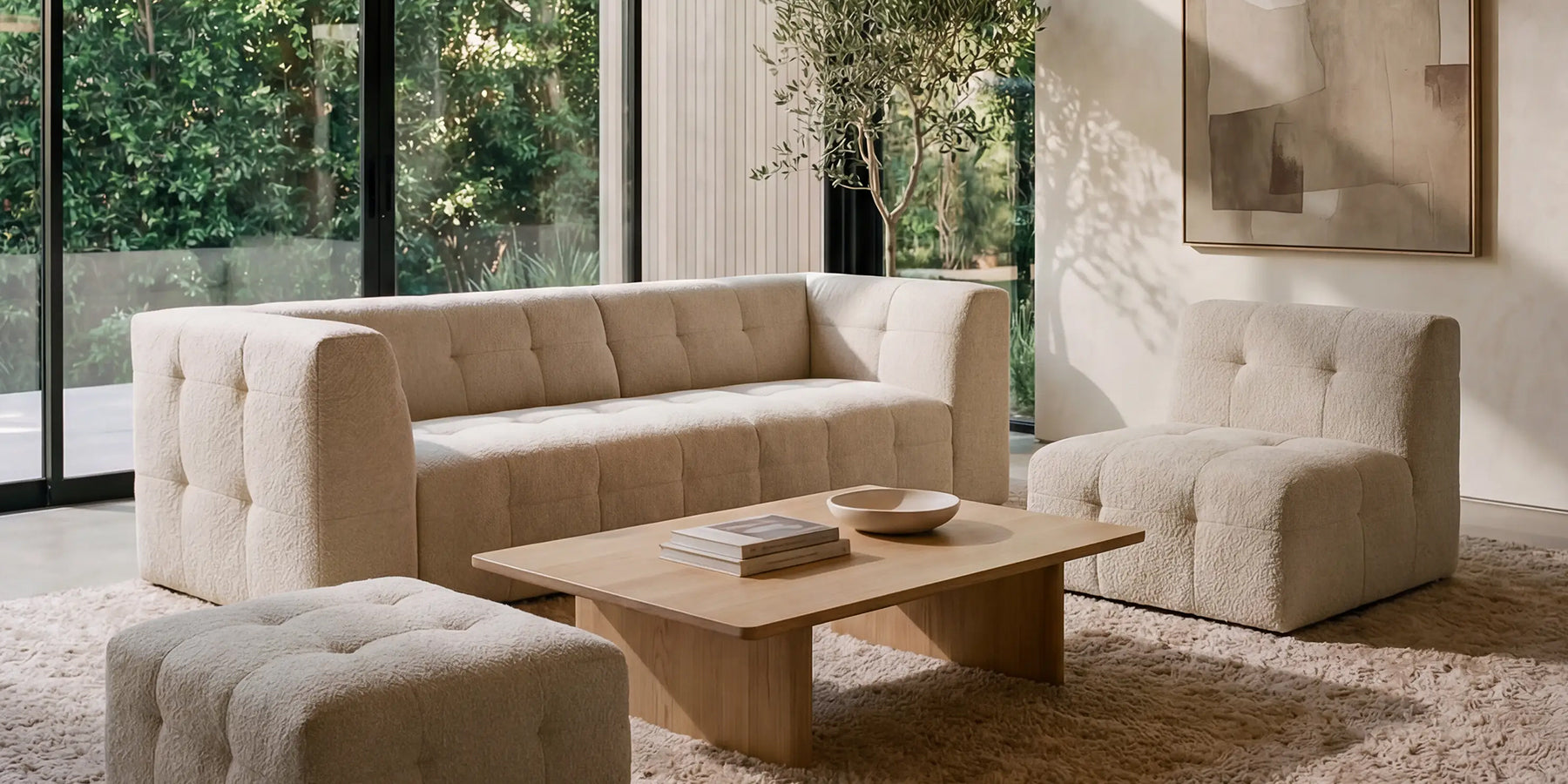 Soggiorno luminoso con un divano trapuntato color crema, due sedie abbinate, un tavolino in legno chiaro con libri e una ciotola, un tappeto morbido, ampie finestre, verde all'esterno, opere d'arte a parete e un'alta pianta da interno.