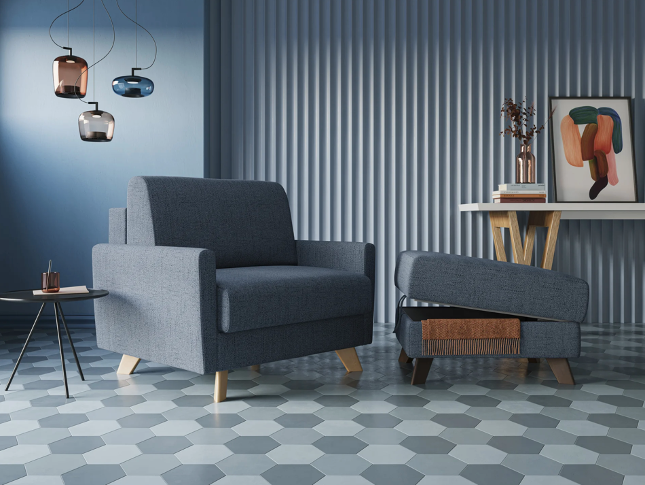 Una moderna poltrona blu e il pouf abbinato sono posizionati su un pavimento in piastrelle esagonali, accanto a un tavolino e una consolle con opere d'arte astratte, vasi e lampade a sospensione, il tutto contro una parete rivestita di pannelli blu.