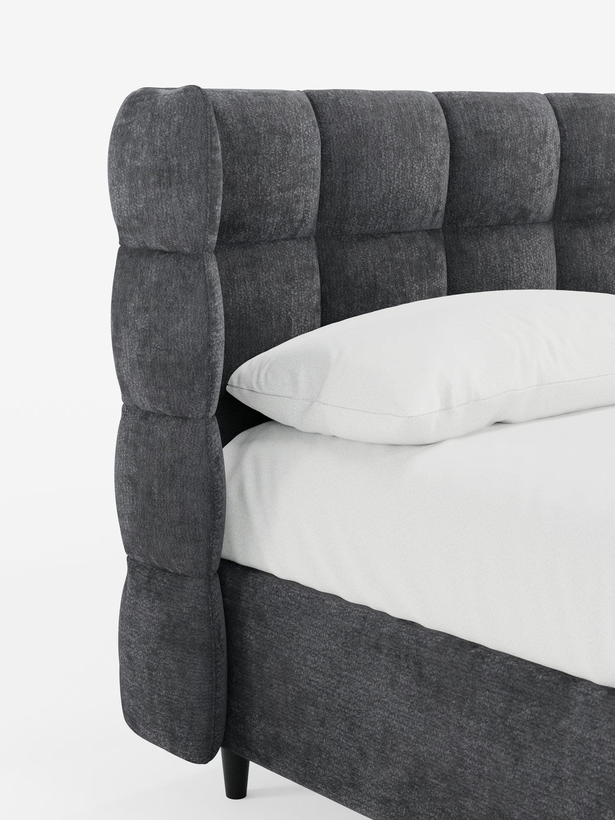 Primo piano del letto imbottito RAGNAR con contenitore e testiera capitonné avvolgente di Letti.store, caratterizzato da una testiera imbottita grigio scuro con spesse e morbide sezioni quadrate in classico stile capitonné. Un elegante tessuto certificato Oeko-Tex® riveste sia la struttura del letto che la testiera abbinate. Sul letto, su uno sfondo bianco a tinta unita, poggiano un cuscino bianco e un lenzuolo bianco.