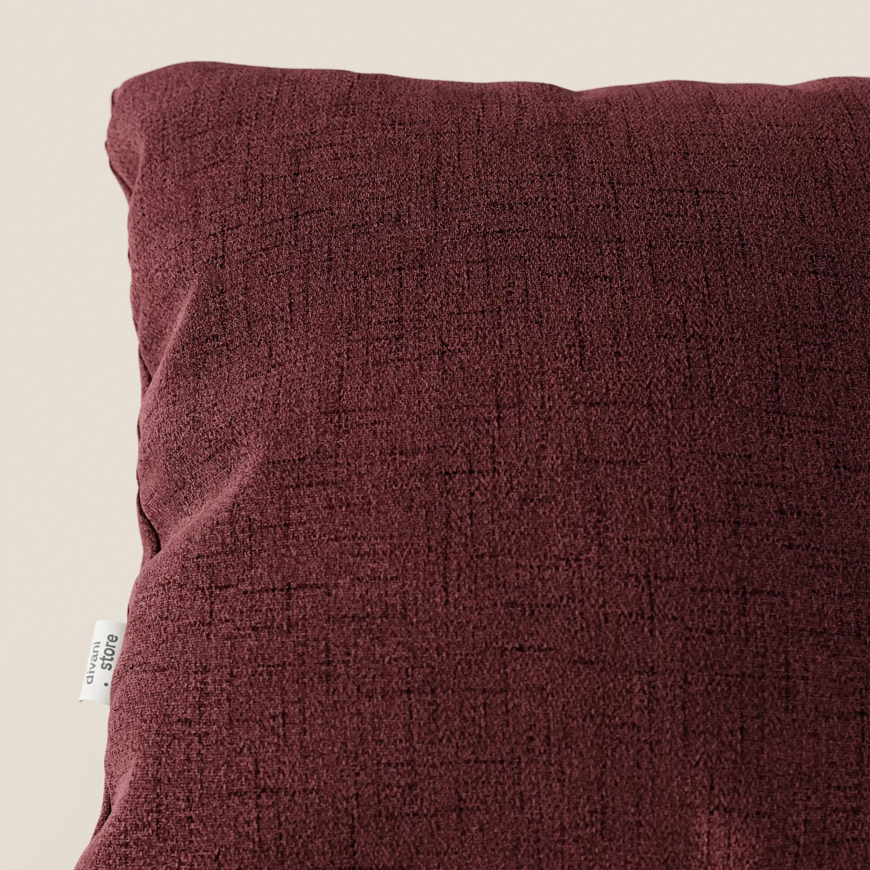 Un primo piano del cuscino Felis cuscini di Divani.Store in T03 rosso, un cuscino quadrato di colore marrone con una sottile trama e un'etichetta in tessuto bianco, realizzato in morbido tessuto impermeabile, su uno sfondo beige chiaro.