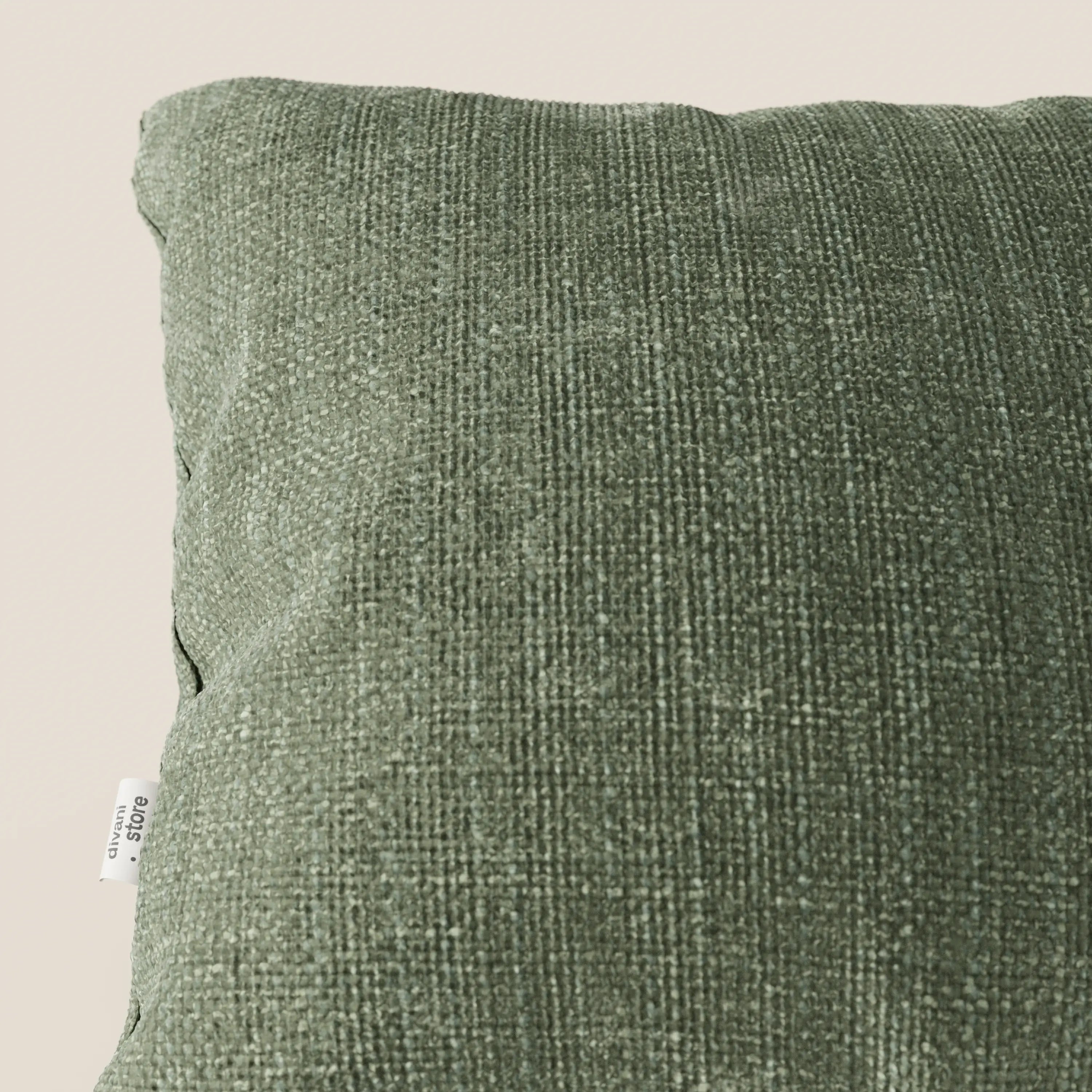 Primo piano del cuscino Lyon 45x45 in tessuto intrecciato impermeabile T06 verde di Divani.Store, caratterizzato da una morbida trama a quadri e da una piccola etichetta in tessuto bianco sulla sinistra, su uno sfondo beige chiaro.
