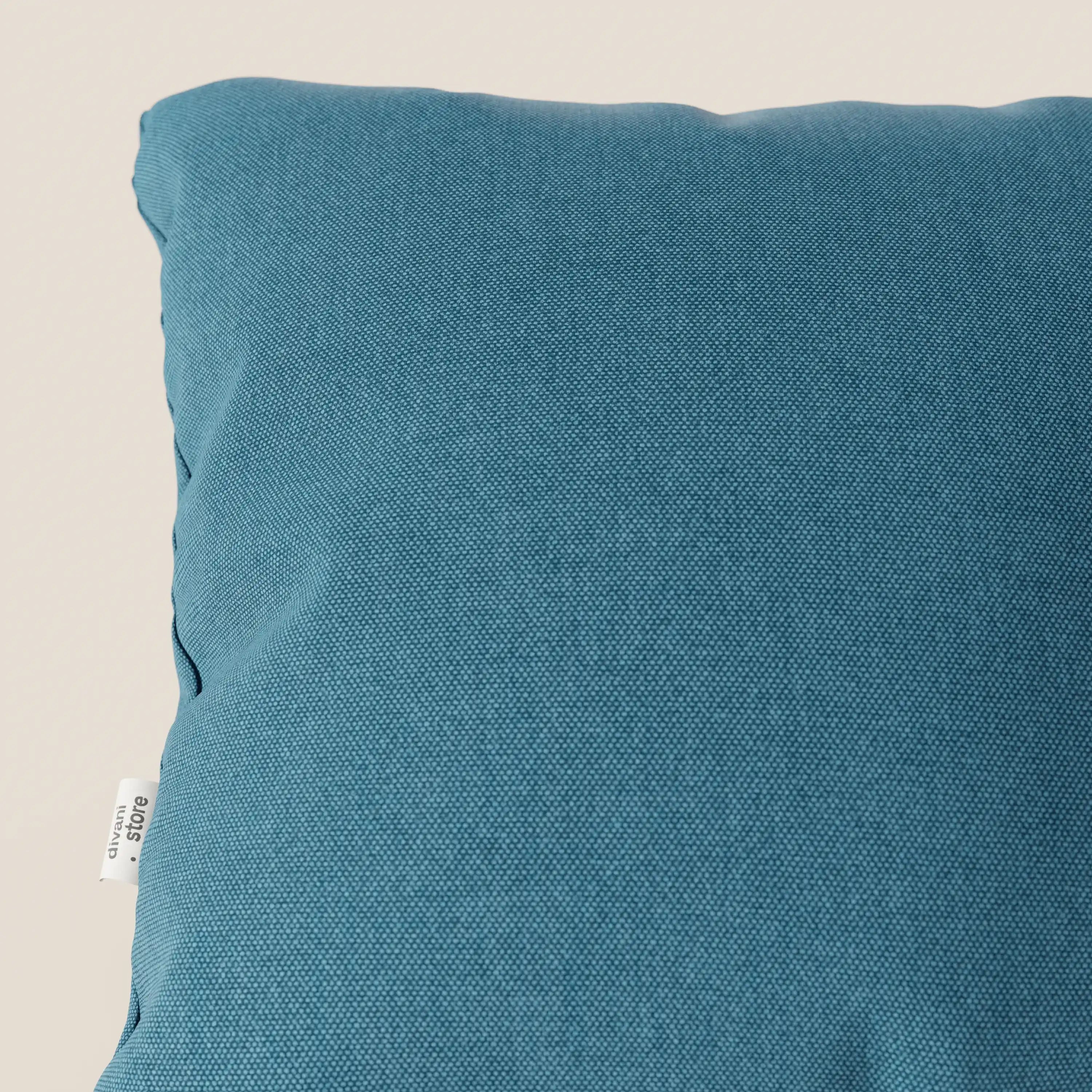 Primo piano del cuscino Honey 45x45 di Divani.Store in T13 azzurro, caratterizzato da un tessuto simil-cotone impermeabile e da una targhetta laterale bianca. Il fondo beige chiaro ne accentua il design elegante.