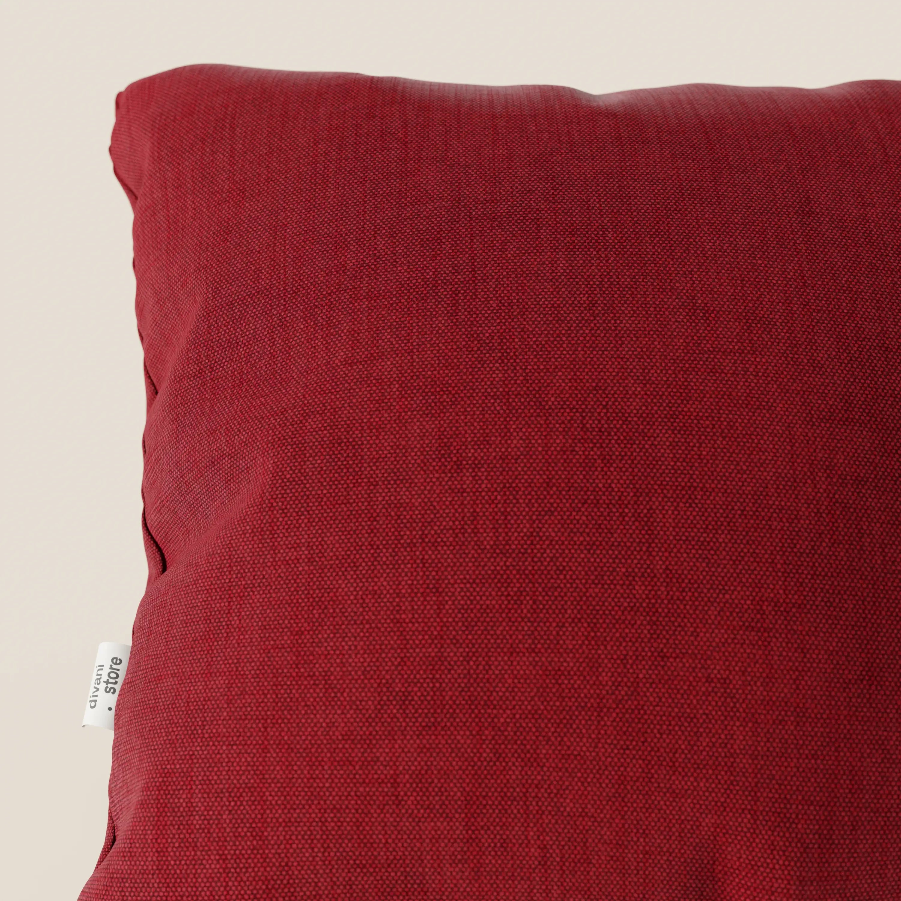 Primo piano del cuscino Divani.Store Honey 45x45 in T13 rosso tessuto simil cotone impermeabile, che presenta un'etichetta bianca e un sottile motivo a fodera effetto cotone su uno sfondo beige chiaro. È visibile solo una parte del cuscino.