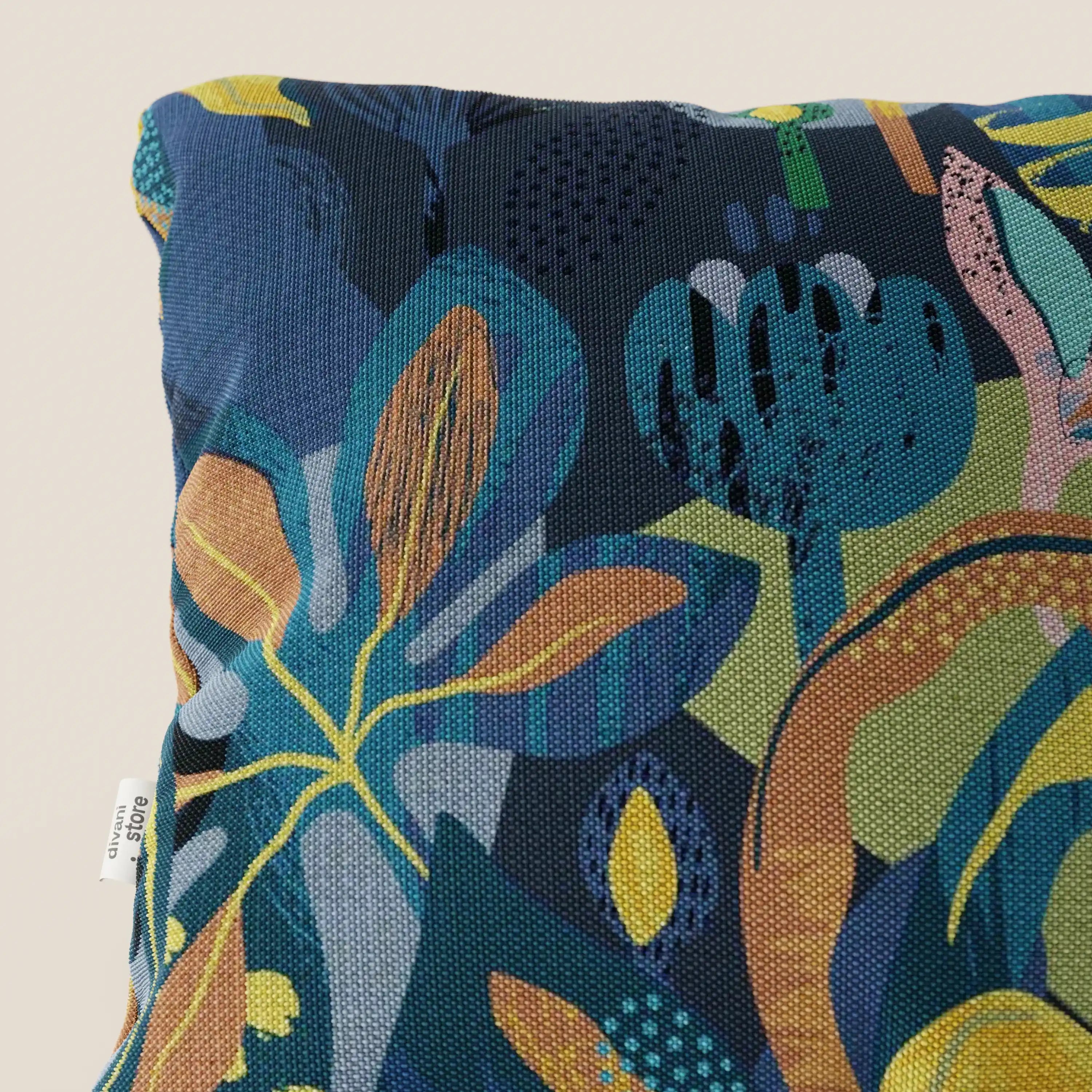 Un primo piano del cuscino Joy di Divani.Store mostra il suo motivo geometrico e botanico multicolore su un fresco cotone blu impermeabile; una piccola etichetta di tessuto bianco appare sulla cucitura sinistra. Lo sfondo è beige.