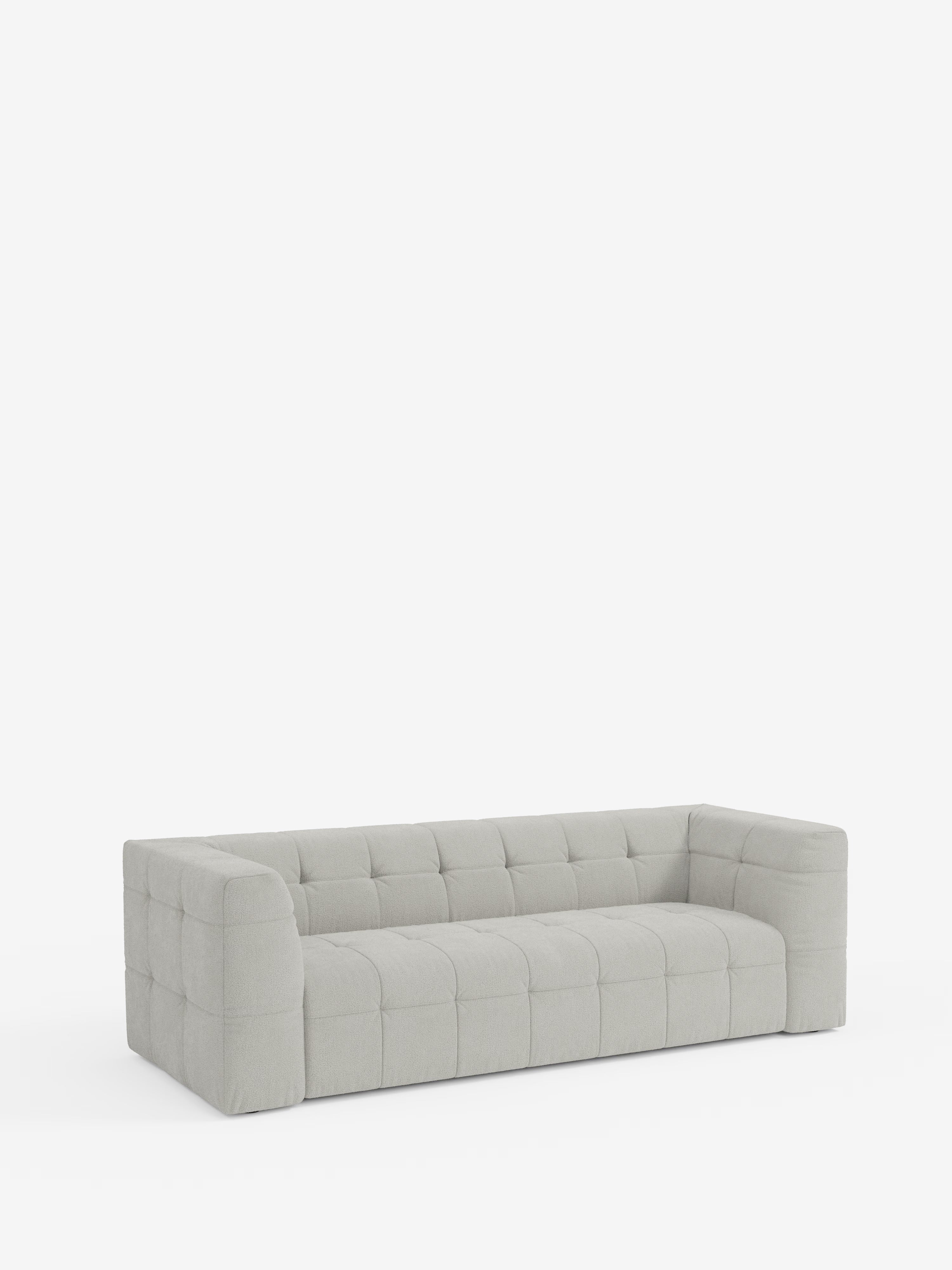 Un divano modulare Mosaic contemporaneo, di colore grigio chiaro, a 3 o 4 posti, di Divani.Store, caratterizzato da ampi braccioli e una seduta profonda. Rivestito in tessuto bouclé T21 antimacchia con trapuntatura rettangolare che crea un effetto a griglia, il design capitonné risalta sullo sfondo bianco a tinta unita, con il divano al centro dell’immagine.