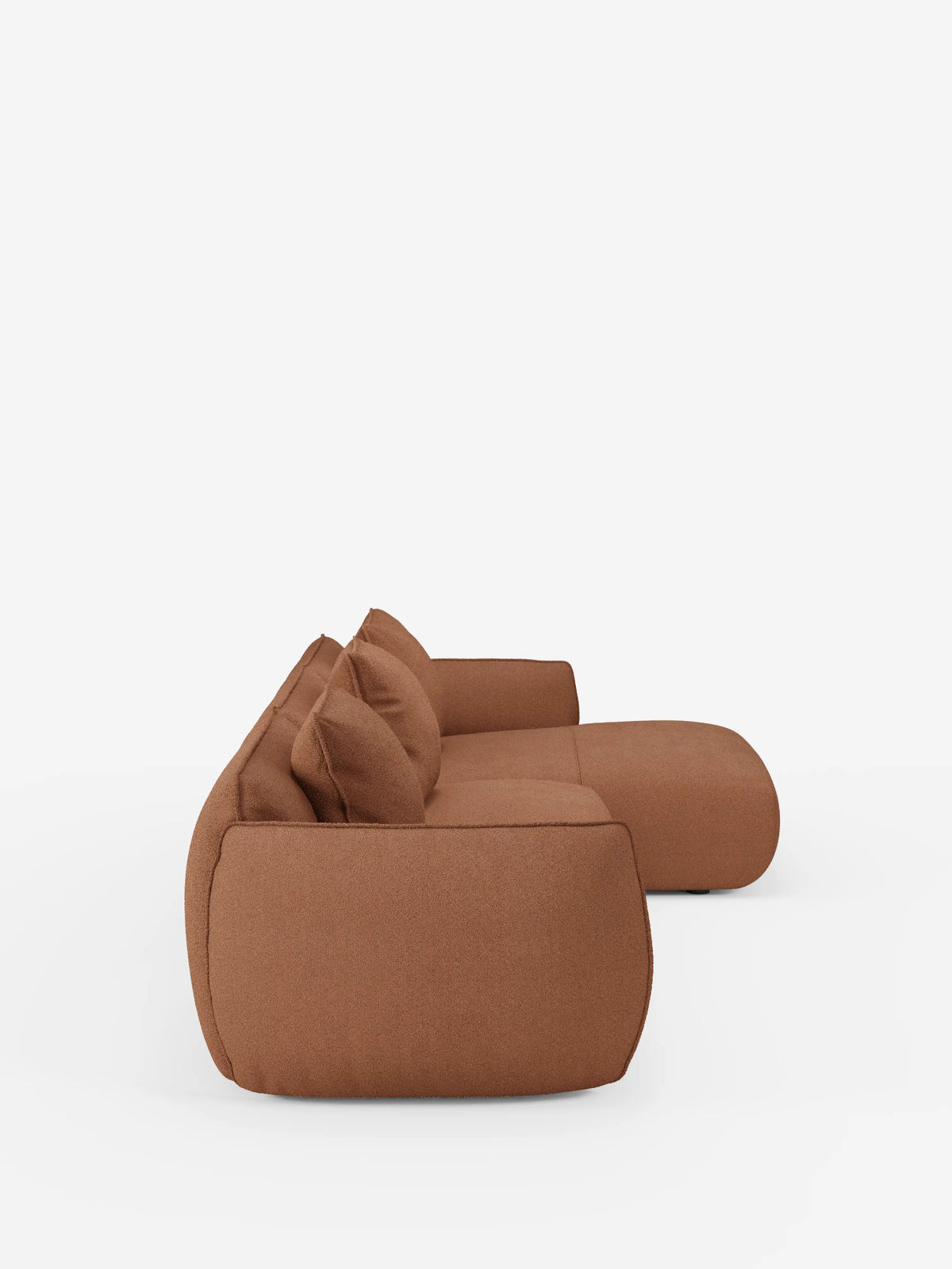Vista laterale del divano angolare con penisola componibile Nimbus in morbido tessuto effetto cotone smacchiabile T33 di Divani.Store, un divano moderno dal profilo basso, di colore marrone, con bordi arrotondati e due cuscini coordinati, in primo piano su uno sfondo bianco a tinta unita. Il suo rivestimento morbido e liscio e il design minimalista conferiscono al divano un aspetto contemporaneo e accogliente.