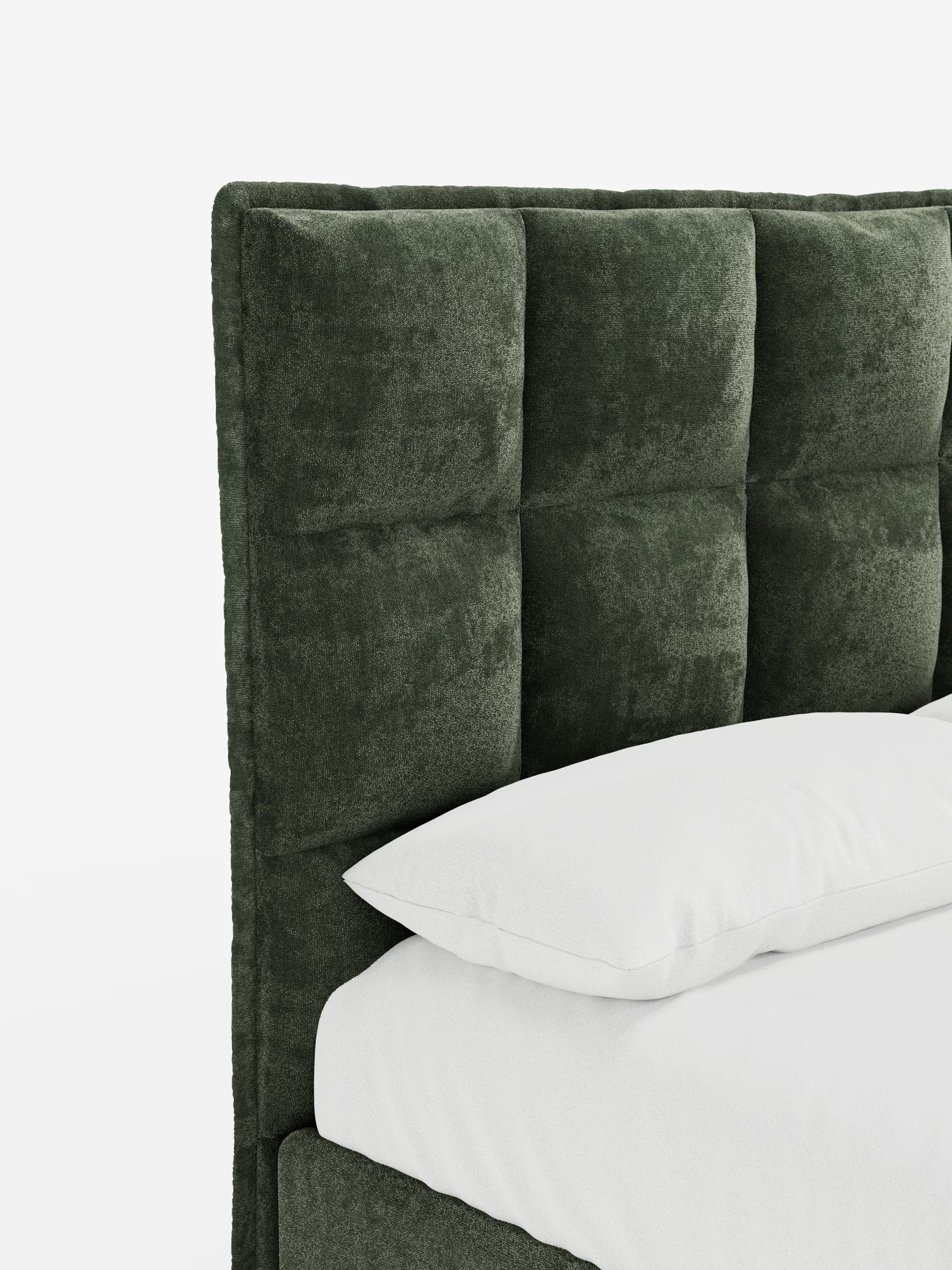 Un primo piano del letto imbottito AURO con contenitore e testiera capitonné alta in ciniglia Oeko-Tex® di Letti.store, caratterizzato da una testiera in tessuto capitonné verde scuro dalla superficie morbida e ruvida al tatto. Il letto è abbellito da un cuscino bianco e da biancheria da letto bianca su uno sfondo bianco, che ne mette in risalto solo una parte della struttura ed esalta il suo design moderno.