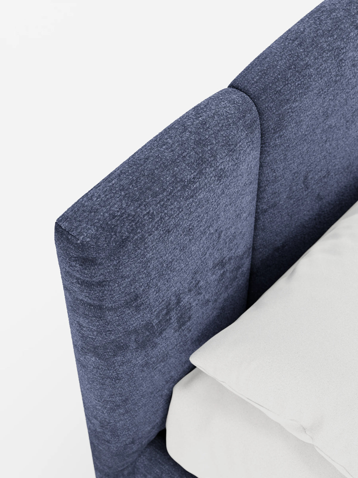 Un primo piano del letto ARKAM imbottito, rivestito in tessuto blu, con contenitore e testiera a lavorazione verticale, in cui spiccano le spesse cuciture verticali accanto a un morbido cuscino bianco su uno sfondo bianco a tinta unita.