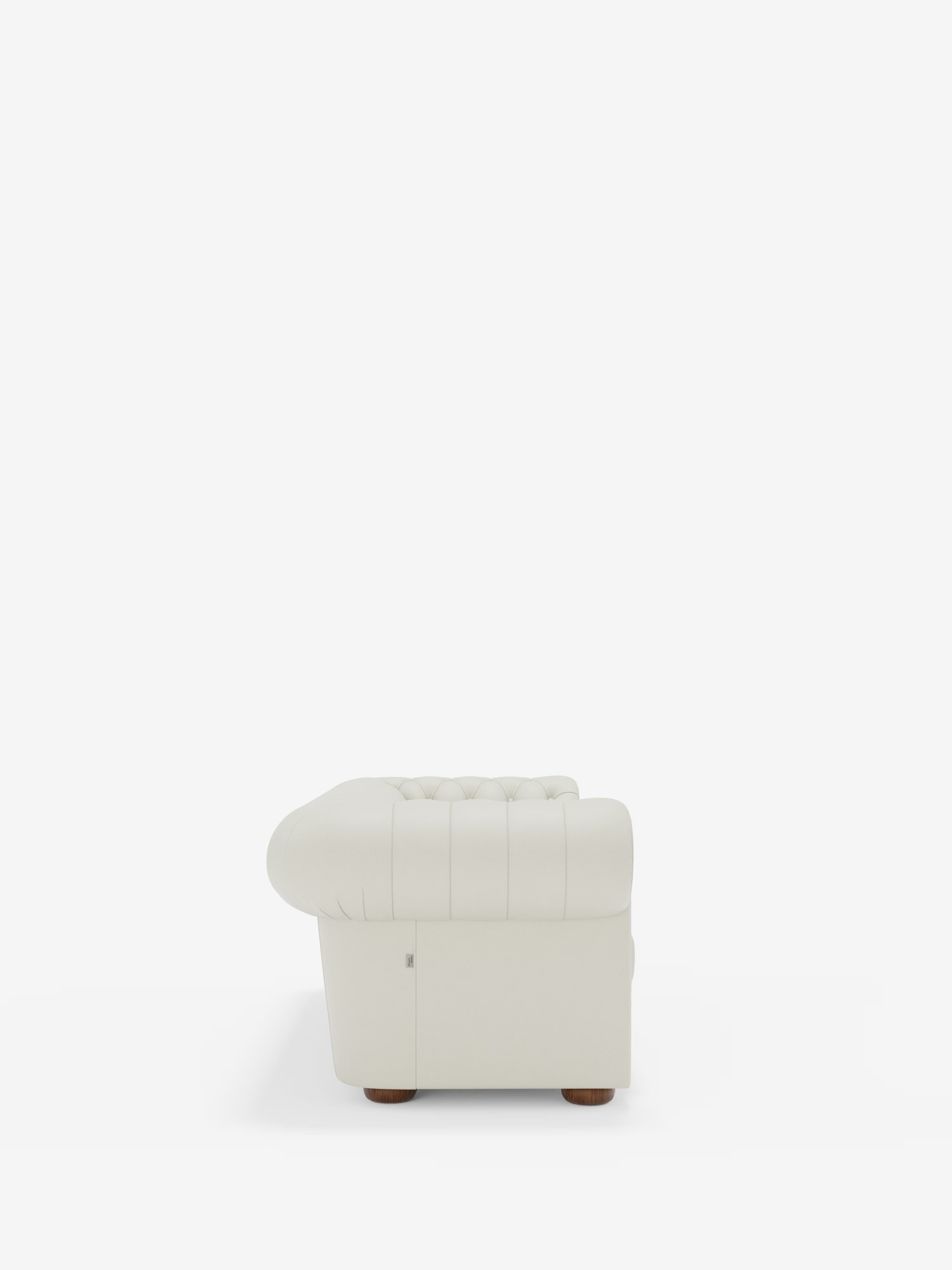 Vista laterale di una poltrona in pelle bianca trapuntata con braccioli arrotondati, cuciture verticali e gambe corte in legno — che richiama il classico stile Chesterfield — su uno sfondo bianco a tinta unita. Il modello Chester – Divano Chesterfield in vera pelle spessorata a 2 e 3 posti di Divani.Store è un esempio di squisita maestria artigianale italiana ed eleganza senza tempo.