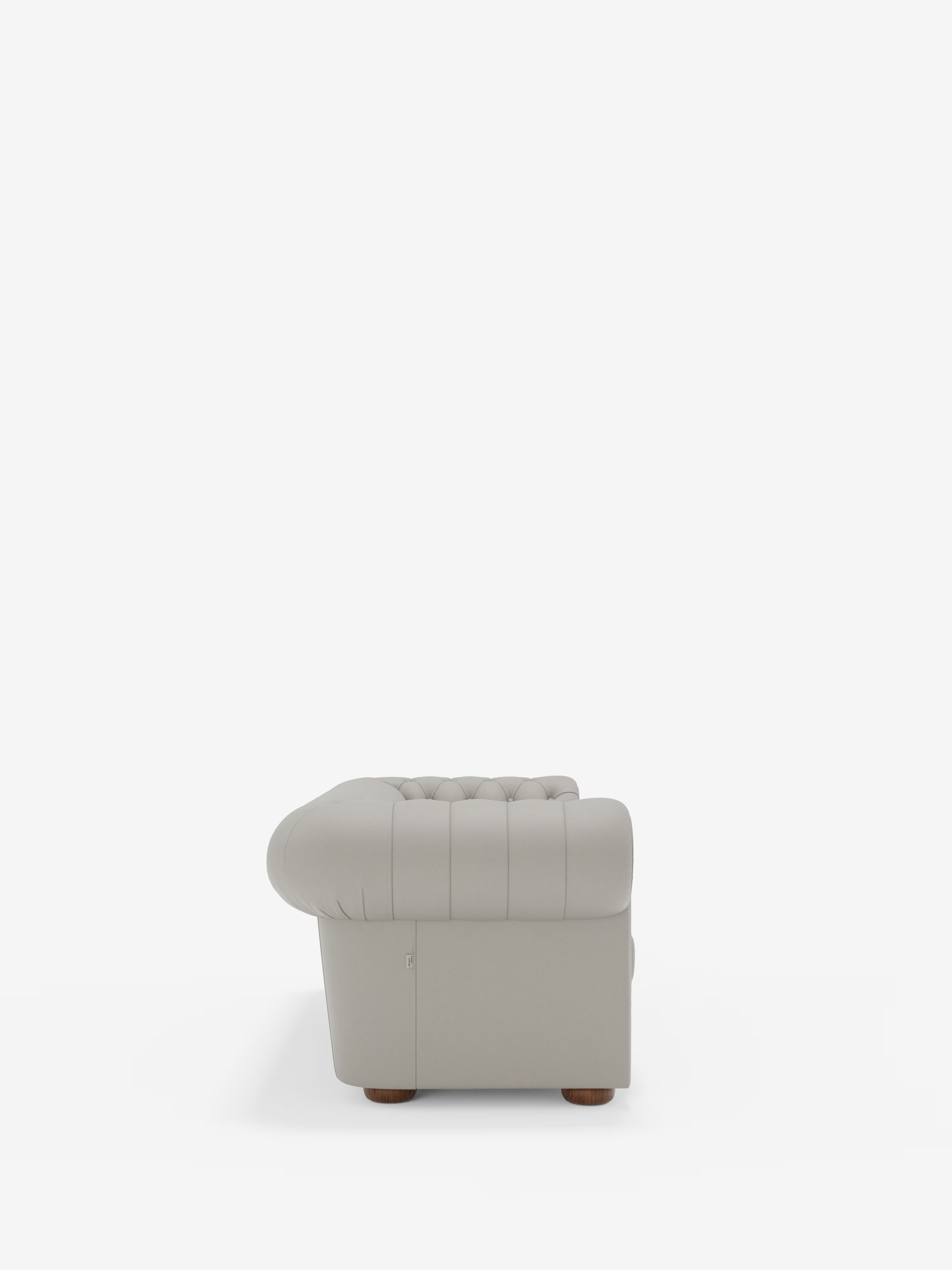 Una vista laterale del modello Chester – Divano Chesterfield in vera pelle spessorata a 2 e 3 posti di Divani.Store, presentato in vera pelle grigio chiaro con rivestimento trapuntato, braccioli arrotondati e robuste gambe in legno su uno sfondo bianco a tinta unita. In linea con l’intramontabile stile Chesterfield, vengono messi in risalto la morbida texture della pelle e i dettagli classici della poltrona, con particolare attenzione alla larghezza e alla maestria artigianale dei braccioli.