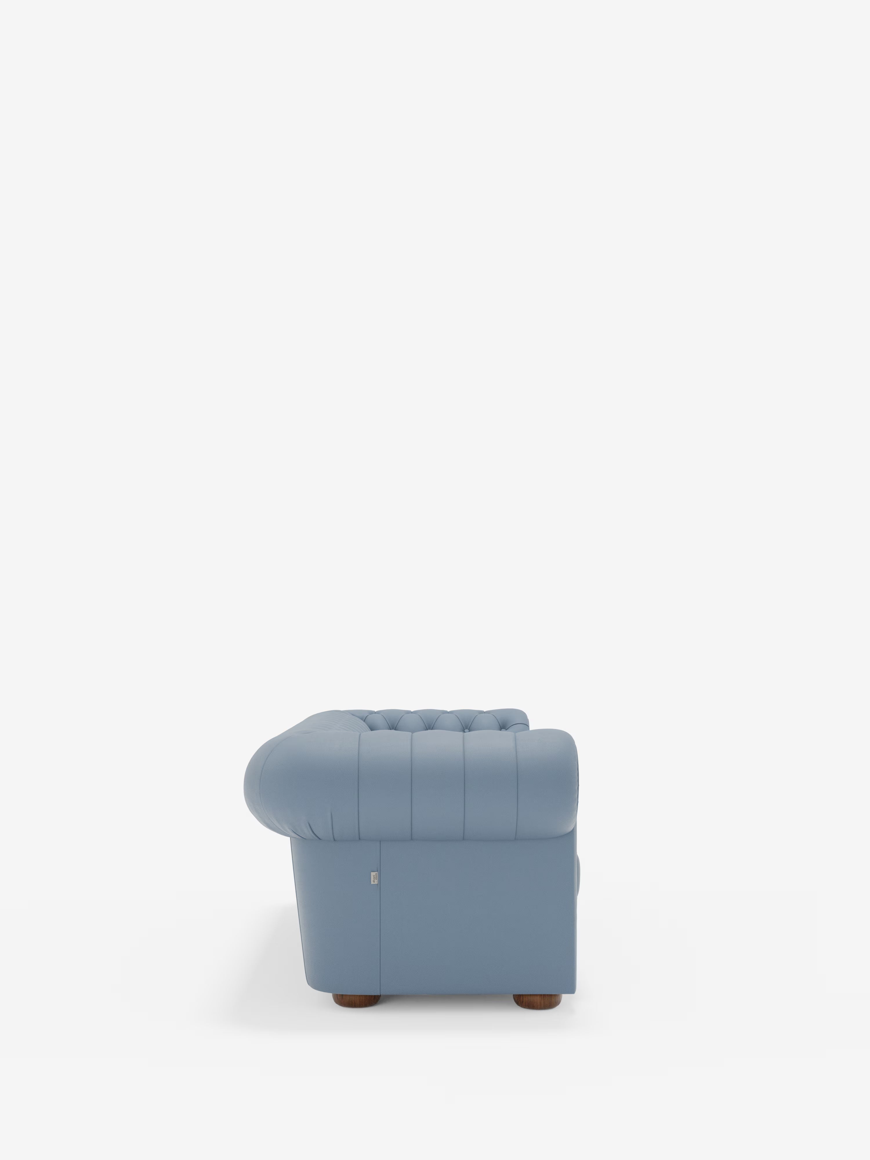 Vista laterale del Chester – Divano Chesterfield in vera pelle spessorata a 2 e 3 posti di Divani.Store, presentato in azzurro con rivestimento trapuntato, braccioli ricurvi e gambe basse in legno su uno sfondo bianco a tinta unita. Questo elegante divano artigianale coniuga un design classico con una raffinata maestria artigianale.