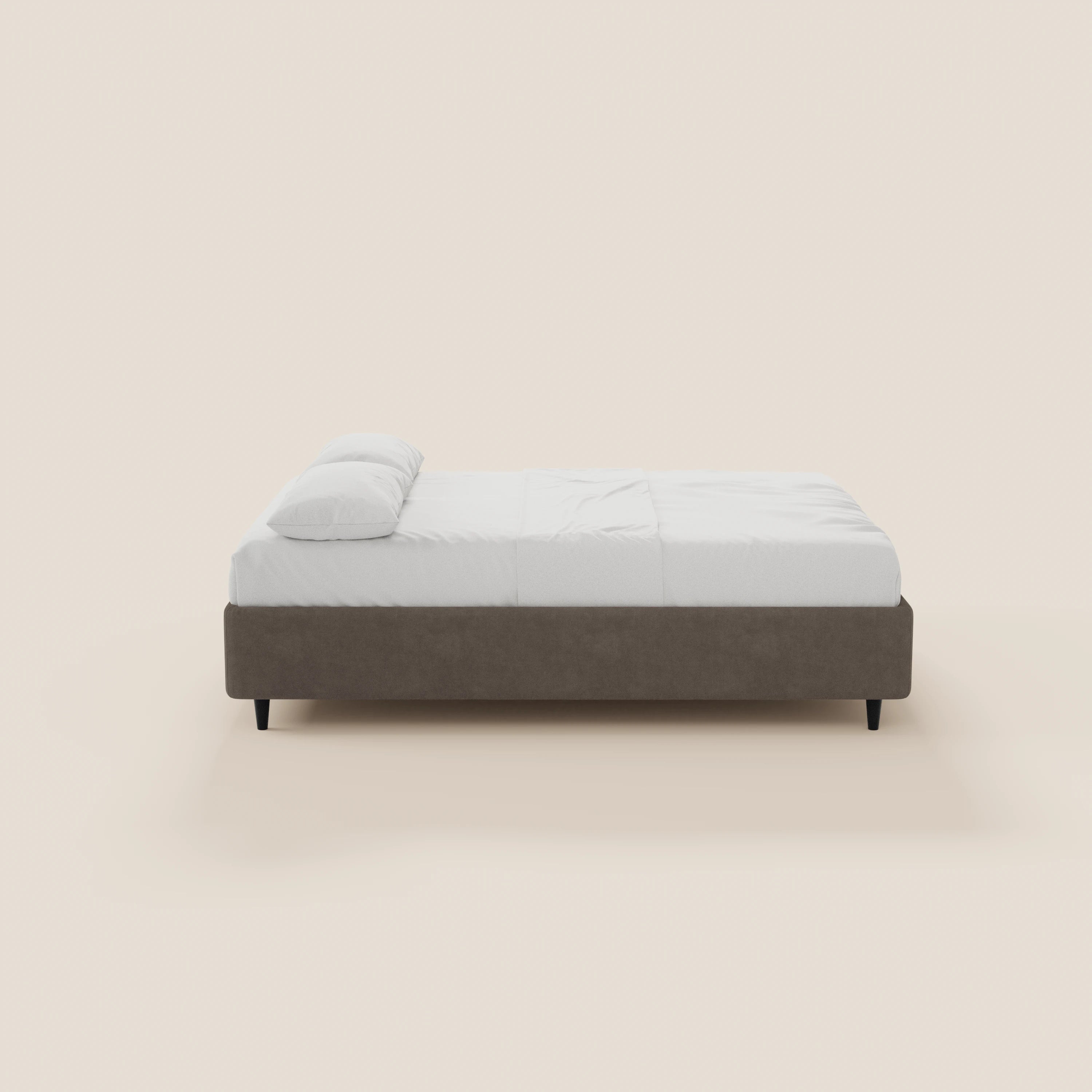 Vista laterale del letto sommier imbottito ERON con vano contenitore in tessuto impermeabile certificato Oeko-Tex di Letti.store, caratterizzato da una struttura marrone chiaro, materasso e biancheria da letto bianchi, gambe nere e un design minimalista su uno sfondo beige.