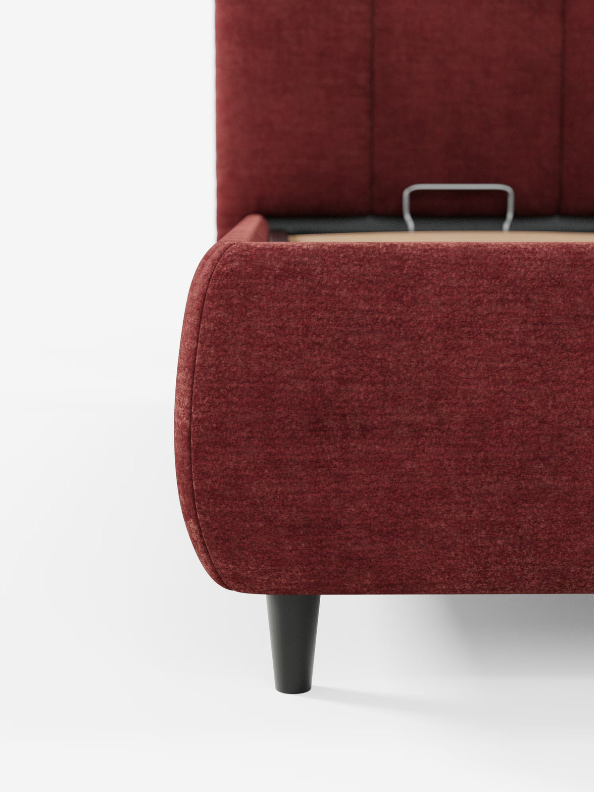 Primo piano dell'angolo inferiore del letto ARKAM imbottito con contenitore e testiera a lavorazione verticale di Letti.store, in rosso, con gambe nere. Il letto presenta un design morbido e sinuoso in stile shabby chic, realizzato in tessuto certificato Oeko-Tex®, e include un cassetto aperto — tipico di un letto contenitore — con una maniglia in metallo parzialmente visibile sul lato. Lo sfondo è bianco a tinta unita.