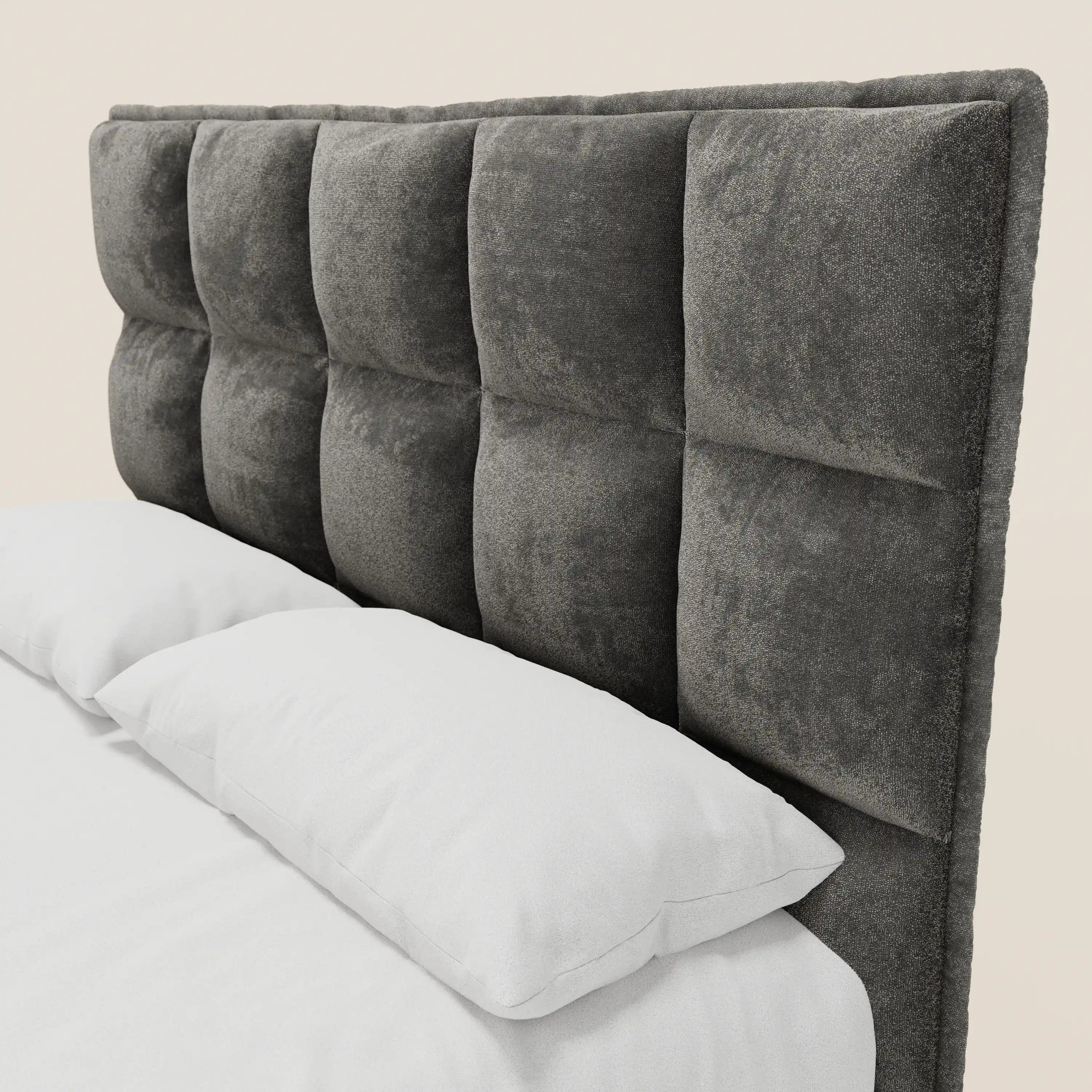 Un primo piano del letto imbottito AURO di Letti.store mostra una testiera grigia in ciniglia con capitonné alto certificata Oeko-Tex®, caratterizzata da ampi pannelli quadrati, biancheria da letto bianca e due cuscini su uno sfondo beige chiaro.