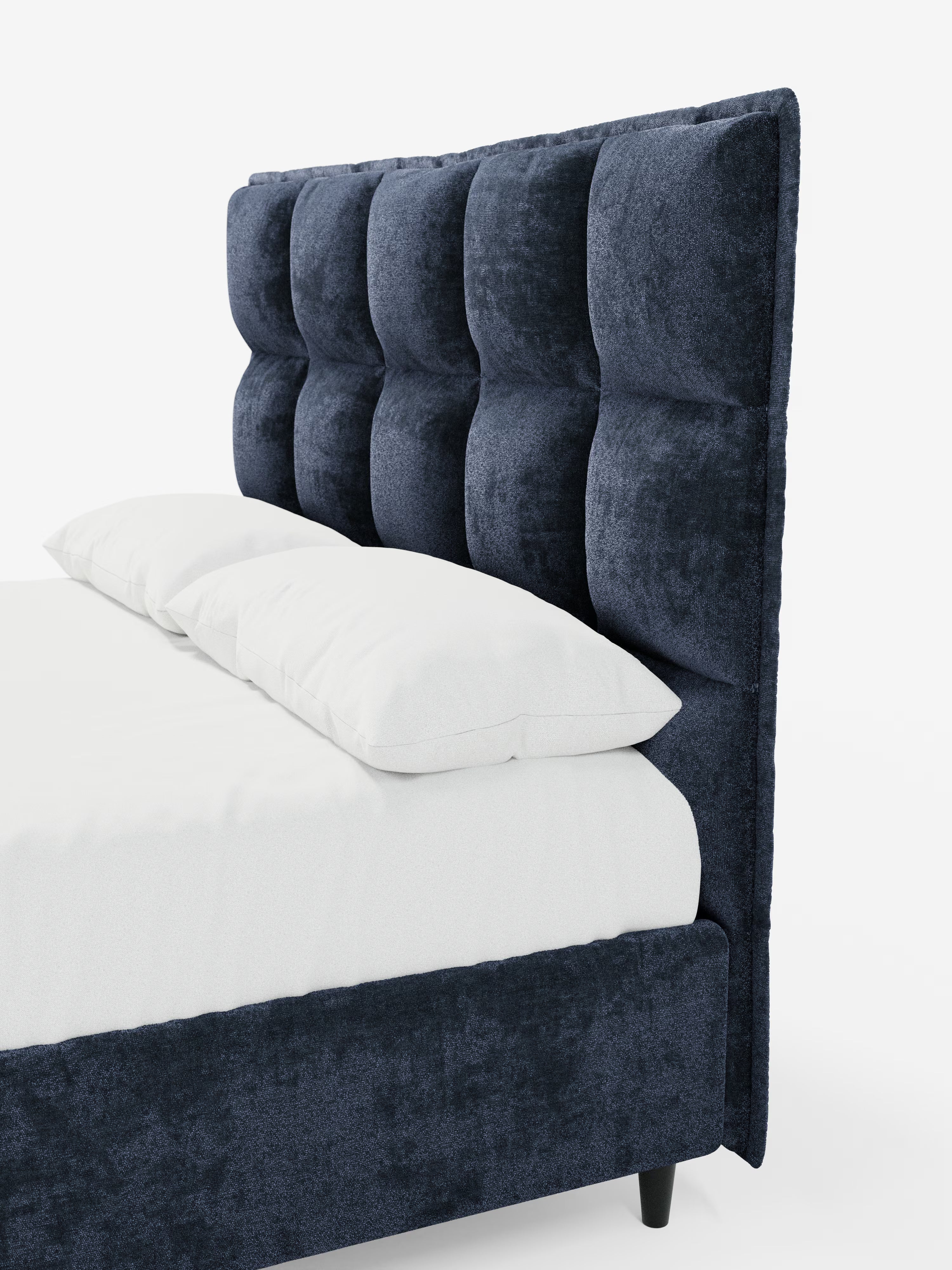 Una vista laterale del letto imbottito AURO con contenitore e testiera alta capitonné in ciniglia Oeko-Tex® di Letti.store, caratterizzato da una testiera alta capitonné in ciniglia blu navy con pannelli rettangolari profondi. Il letto è rivestito con biancheria da letto bianca e due cuscini bianchi e poggia su gambe corte nere su uno sfondo bianco a tinta unita.
