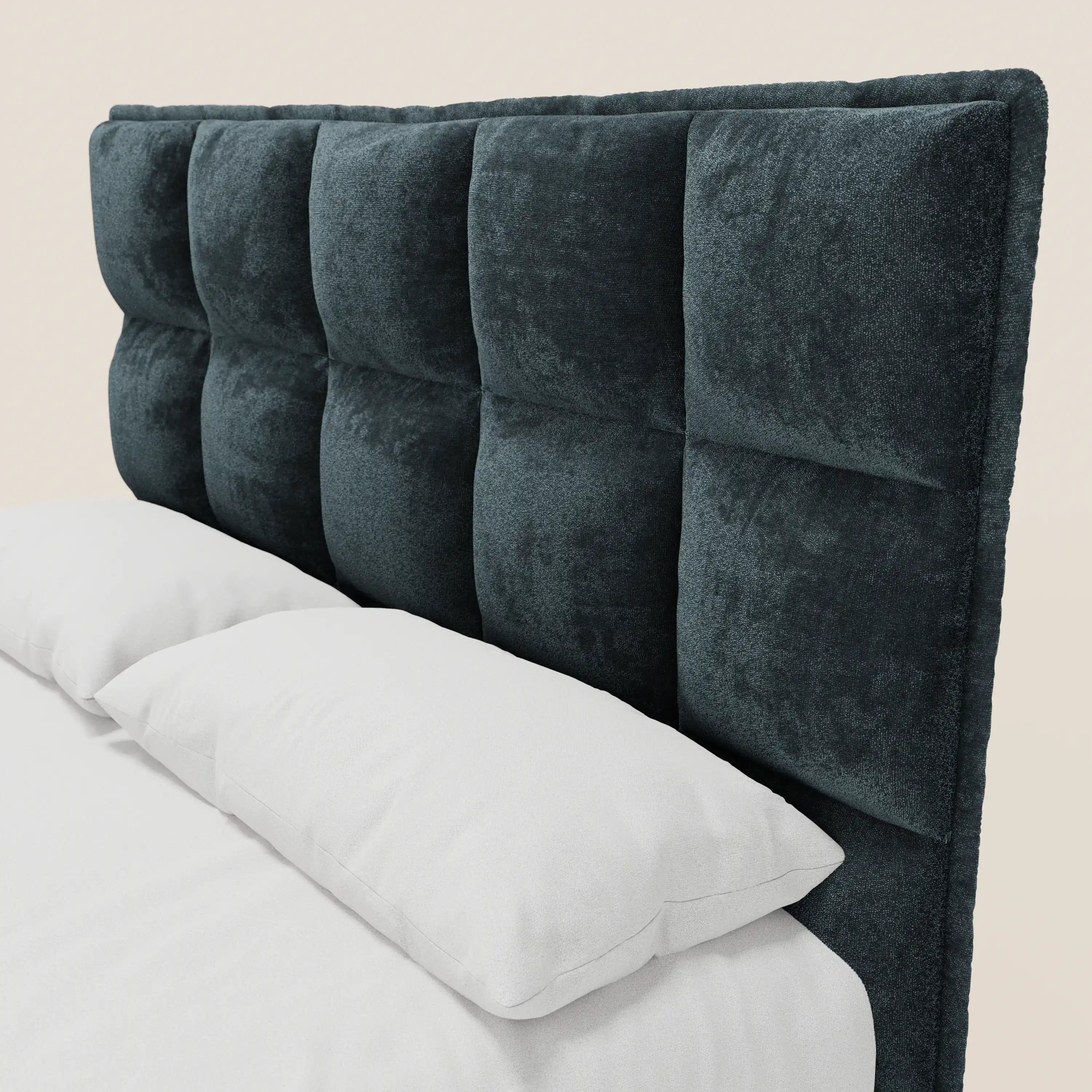 Un primo piano del letto imbottito AURO di Letti.store mette in risalto la sua testiera alta, di colore grigio scuro, con finitura capitonné in ciniglia certificata Oeko-Tex®, caratterizzata da ampi quadrati imbottiti, biancheria da letto e cuscini bianchi, il tutto su uno sfondo chiaro.