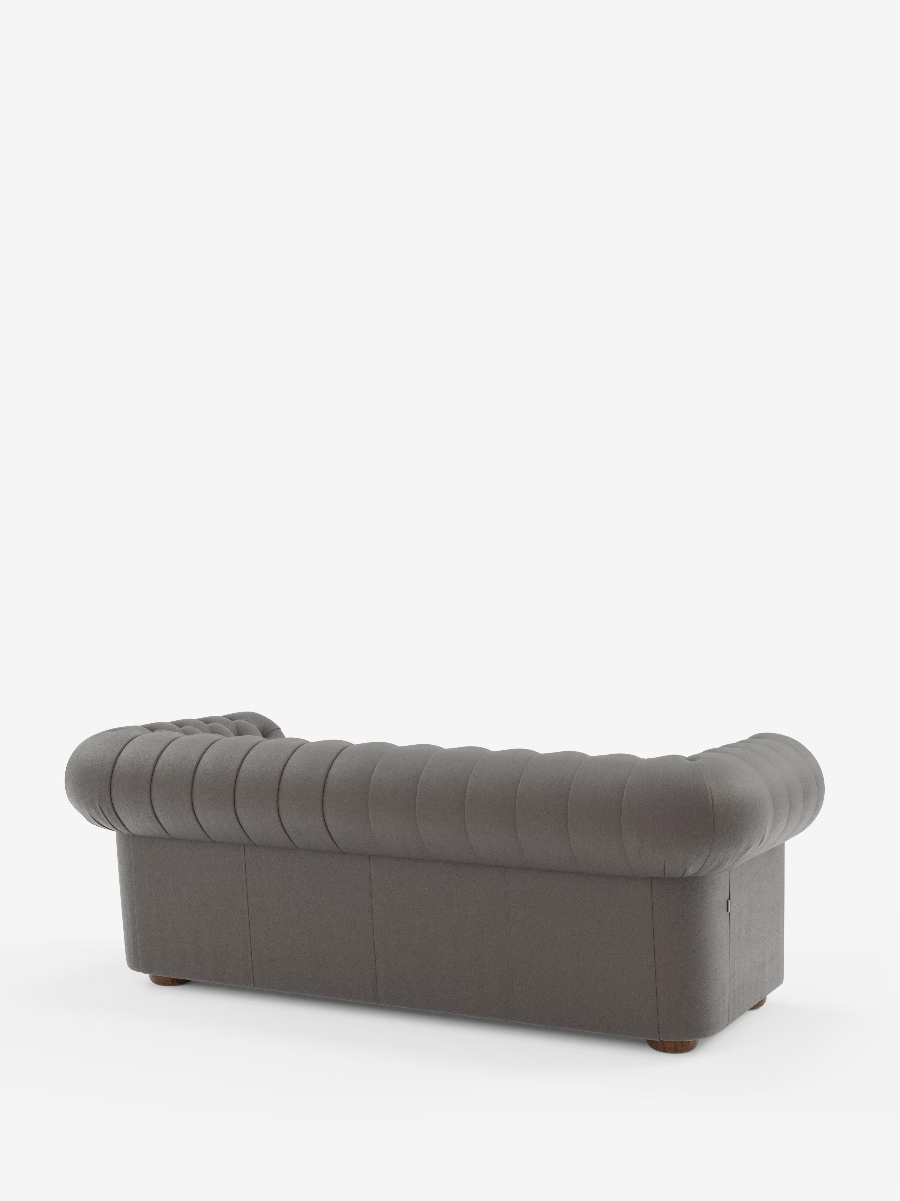 Un divano Chesterfield grigio scuro – Divano Chesterfield in vera pelle spessorata a 2 e 3 posti di Divani.Store – è ripreso da dietro, mettendo in risalto i braccioli ricurvi e arrotondati e lo schienale trapuntato. Questo elegante divano artigianale italiano poggia su piccole gambe rotonde in legno, in primo piano su uno sfondo bianco a tinta unita.